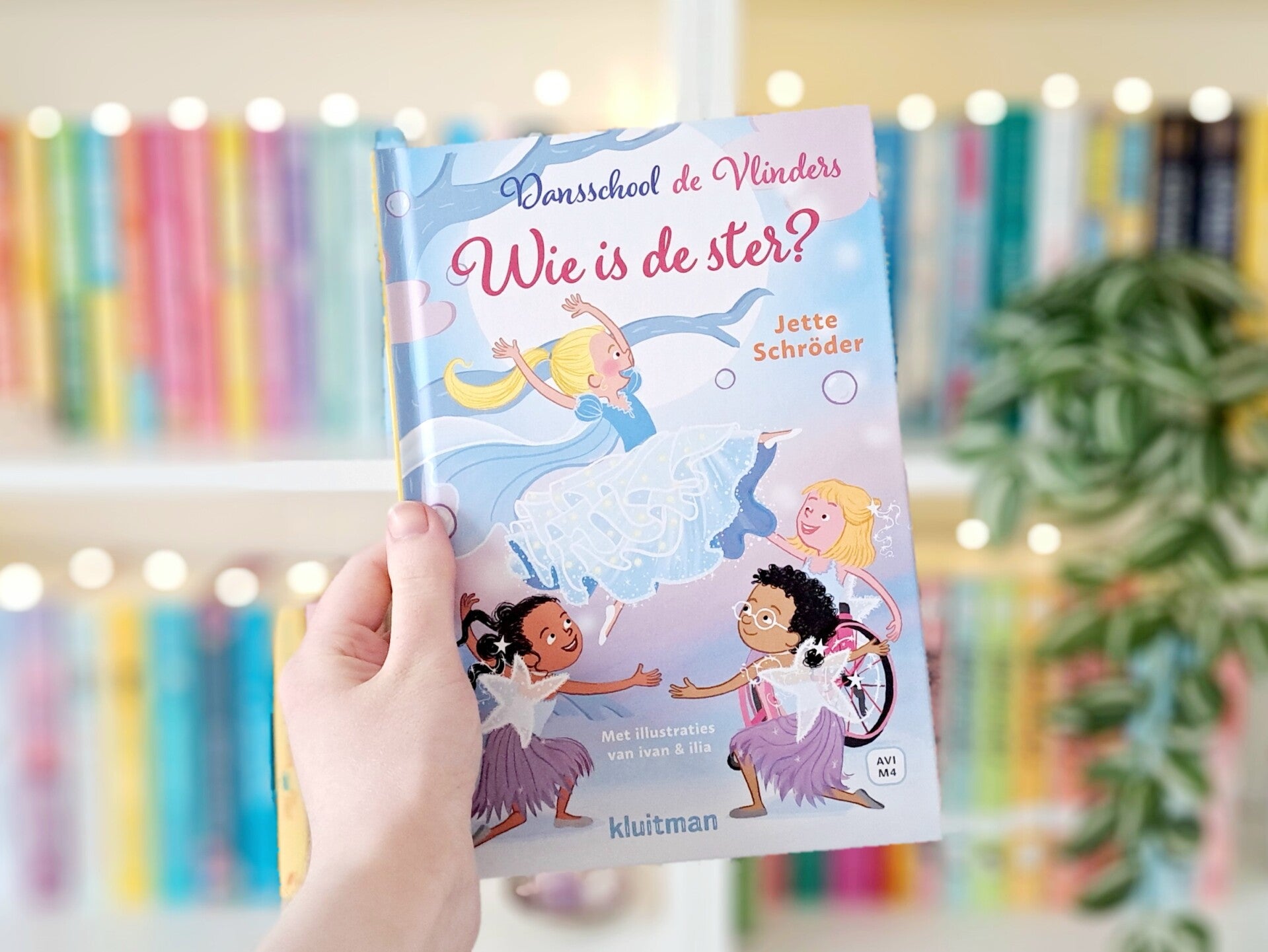 Een witte hand houdt 'Wie is de ster?' deel drie in AVI serie Dansschool de Vlinders van Jette Schröder, geïllustreerd door ivan en ilia, omhoog voor een kleurrijke wazige boekenkast met lichtjes. Foto bij own-voices recensie door turnthepages.nl