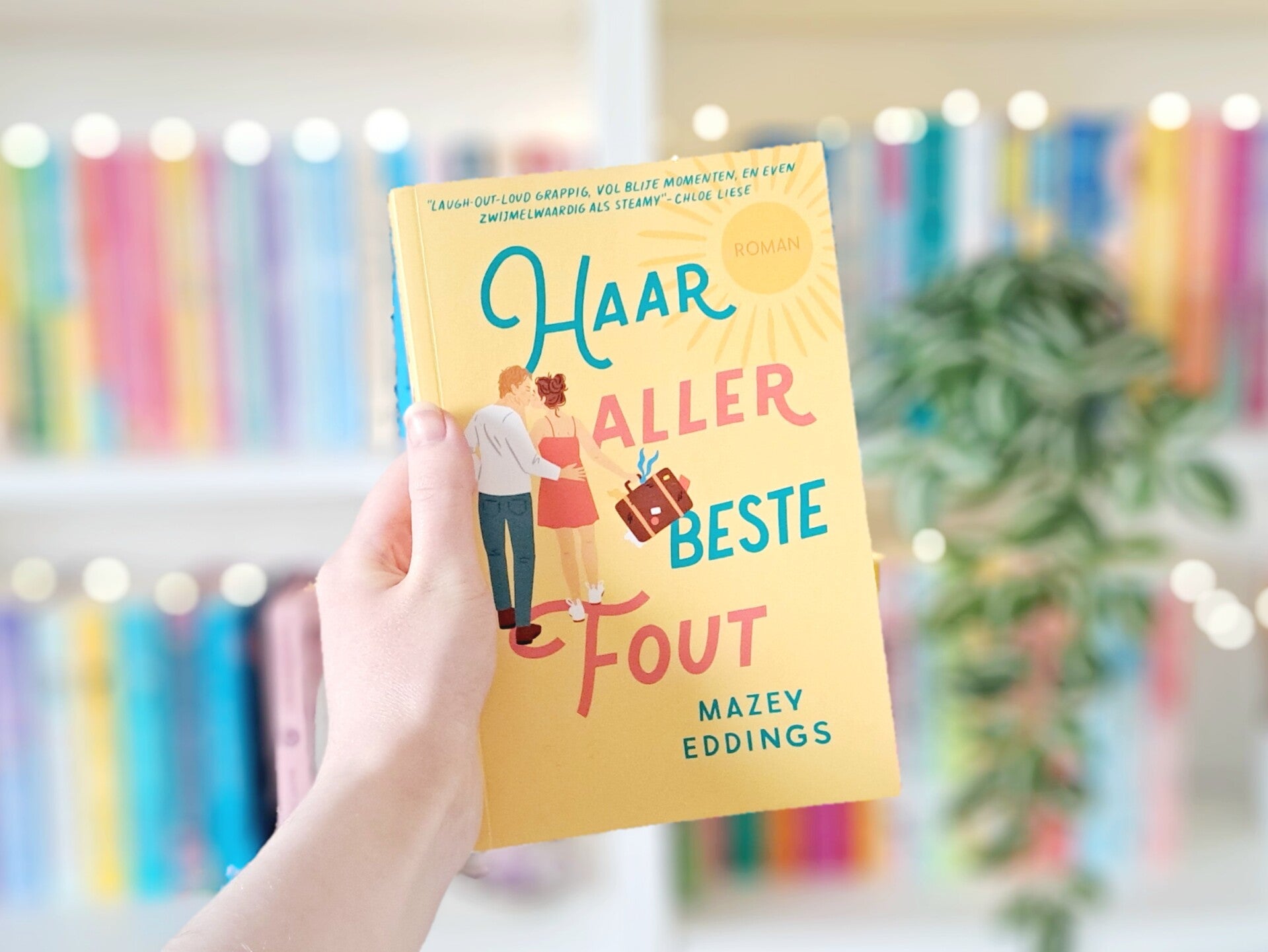 Een witte hand houdt 'Haar allerbeste fout' van Mazey Eddings omhoog voor een kleurrijke wazige boekenkast met lichtjes. Foto bij recensie door turnthepages.nl Rolstoelgebruiker en disabled bookstagrammer en blogger met ADHD.
