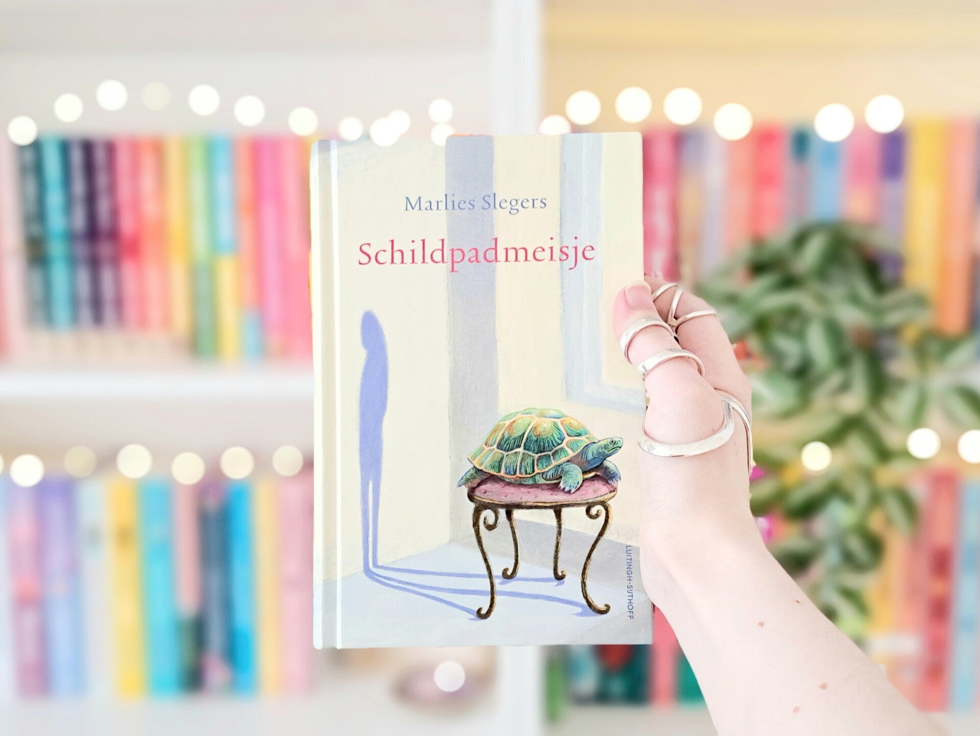 Een witte hand houdt 'Schildpadmeisje' van Marlies Slegers omhoog voor een kleurrijke wazige boekekast met lichtjes. Recensie door turnthepages.nl | Disabled bookstagrammer en neurodivergente boekblogger.