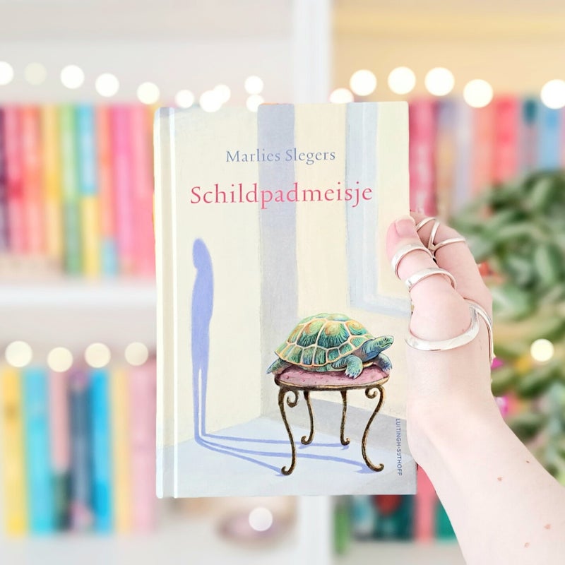 Een witte hand houdt 'Schildpadmeisje' van Marlies Slegers omhoog voor een kleurrijke wazige boekenkast met lichtjes. Foto bij recensie door turnthepages.nl | Rolstoelgebruiker en disabled bookstagrammer en neurodivergent boekblogger.