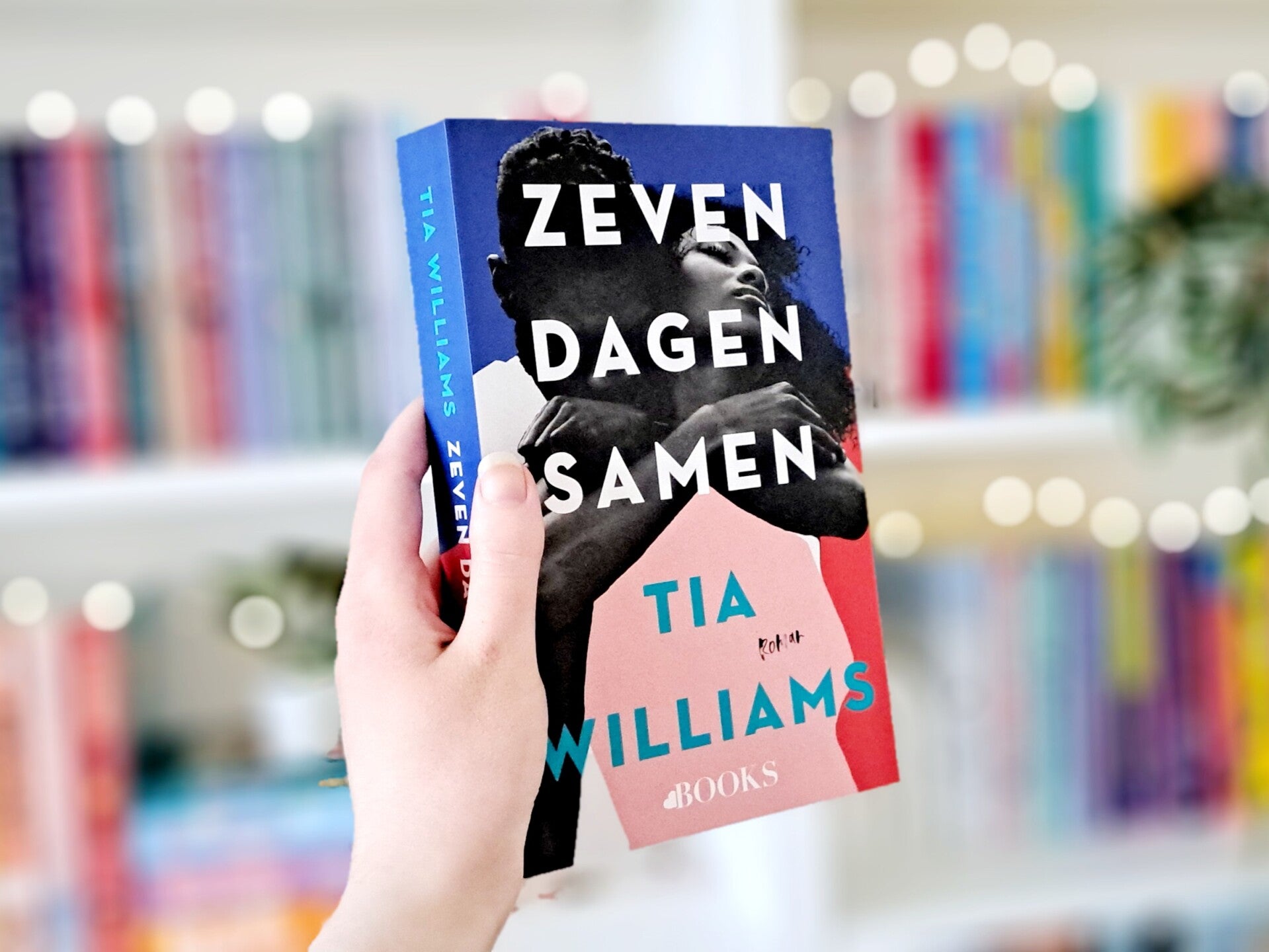 Een witte hand houdt 'Zeven dagen samen' van Tia WIlliams omhoog voor een kleurrijke wazige boekenkast met lichtjes. Recensie door turnthepages.nl Rolstoelgebruiker en disabled bookstagrammer en blogger.