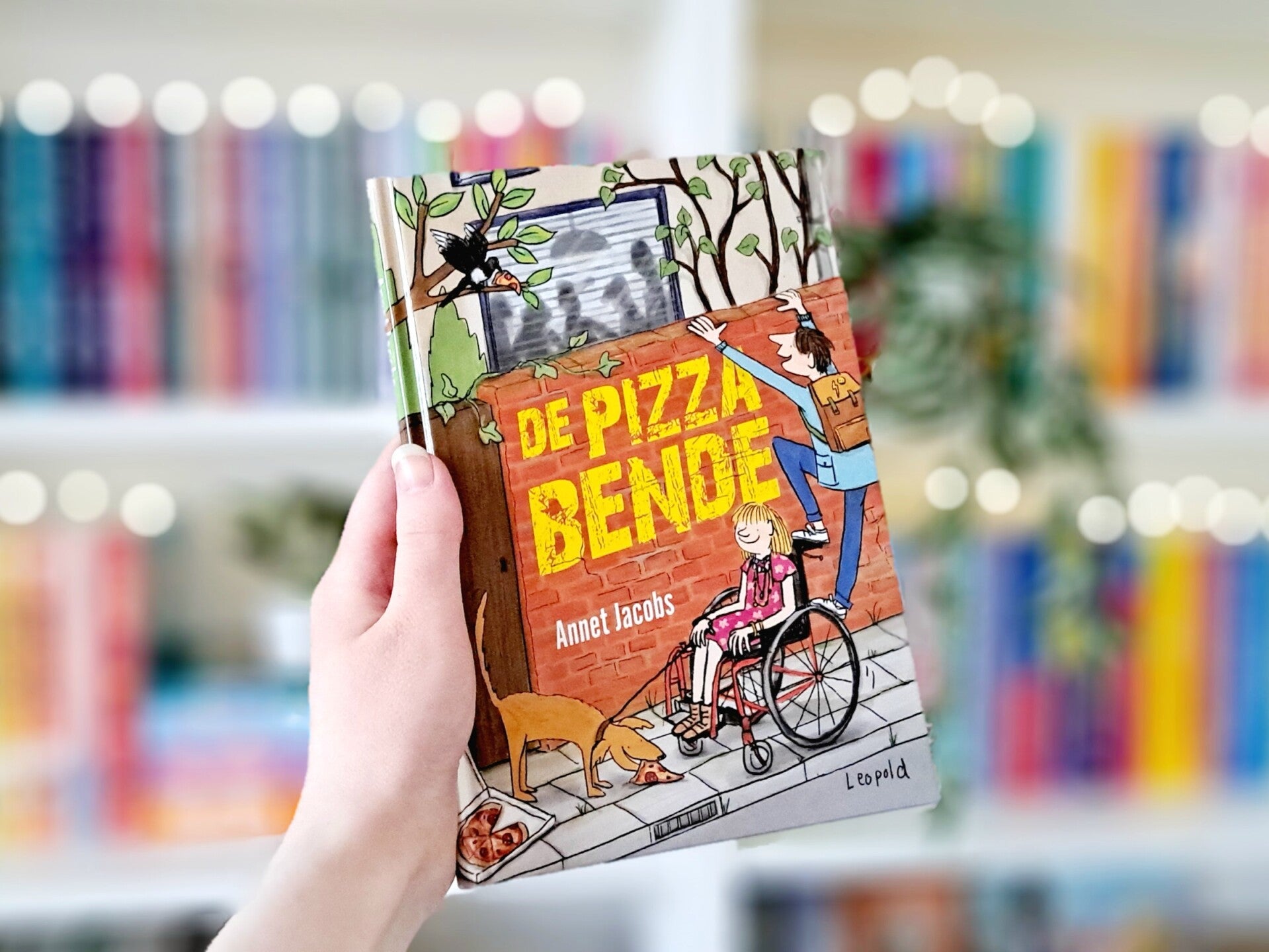 Een witte hand houdt 'De pizzabende' van Annet Jacobs omhoog voor een kleurrijke wazige boekenkast met lichtjes. Foto bij recensie door turnthepages.nl ROlstoelgebruiker en dissabled bookstagrammer en blogger.