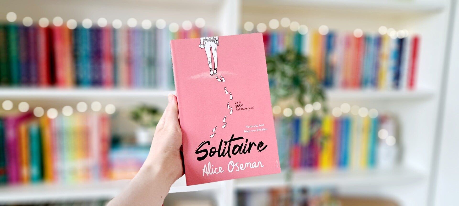 Een witte hand houdt 'Solitaire' van Alice Oseman omhoog voor een kleurrijke wazige boekenkast met lichtjes. Foto bij recensie door turnthepages.nl Disabled bookstagrammer en queer blogger.