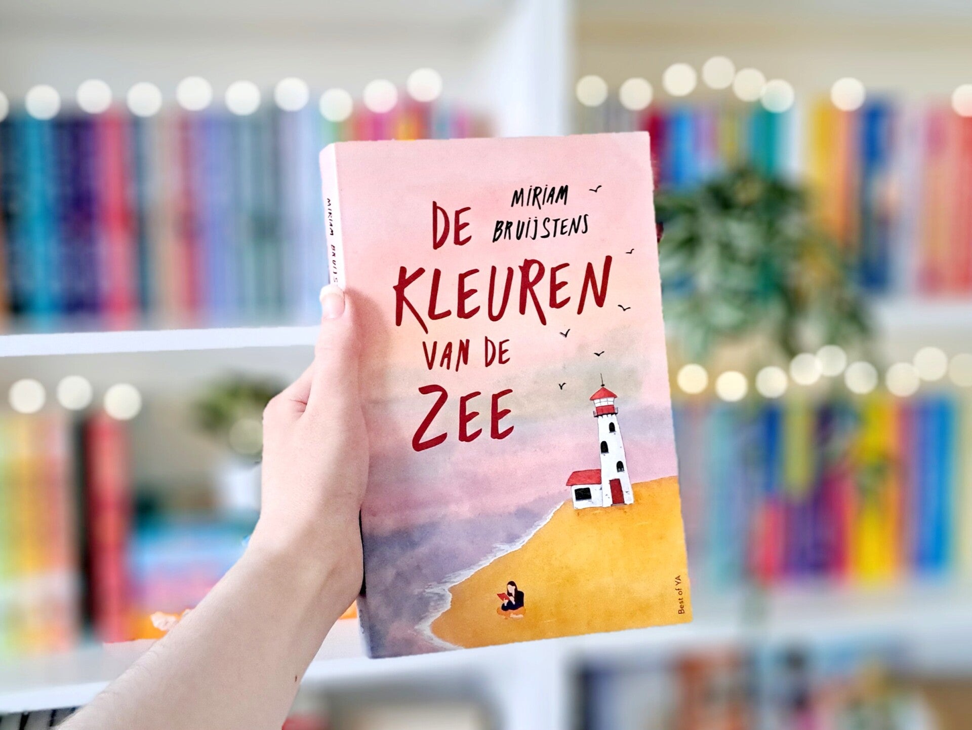 Een witte hand houdt 'De kleuren van de zee' van Miriam Bruijstens omhoog voor een kleurrijke wazige boekenkast met lichtjes. Foto bij own-voices recensie door turnthepages.nl Rolstoelgebruiker en disabled bookstagrammer en blogger met ADHD.