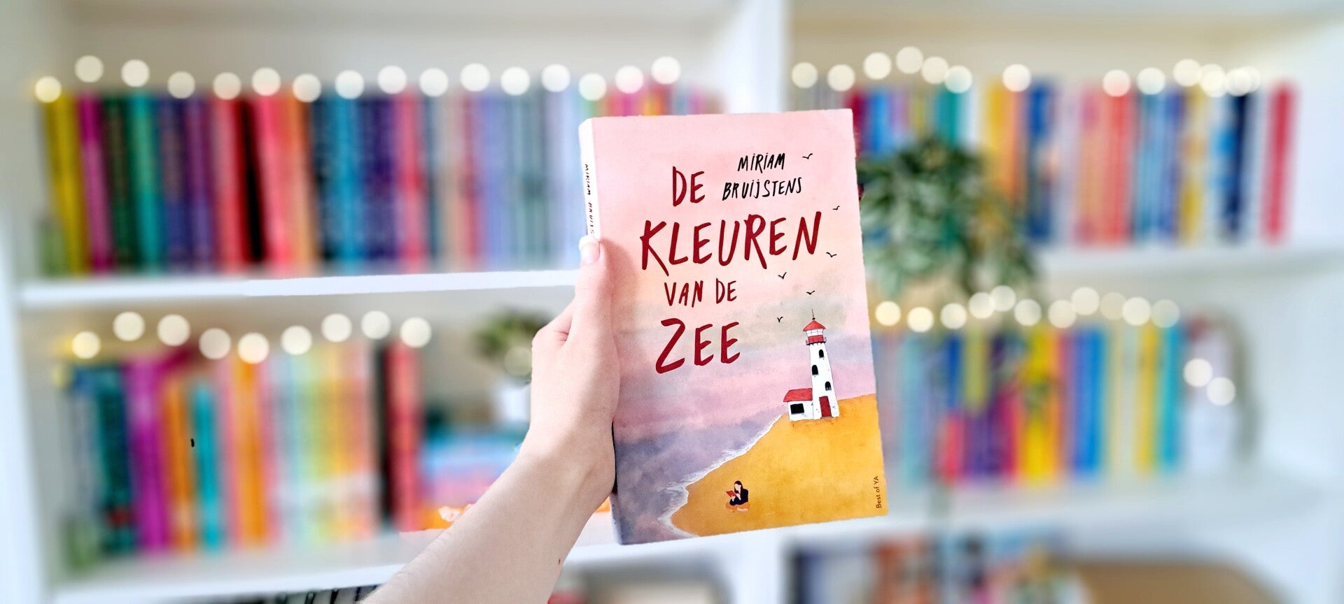 Een witte hand houdt 'De kleuren van de zee' van Miriam Bruijstens omhoog voor een kleurrijke wazige boekenkast met lichtjes. Foto bij own-voices recensie door turnthepages.nl Rolstoelgebruiker en disabled bookstagrammer en blogger met ADHD.