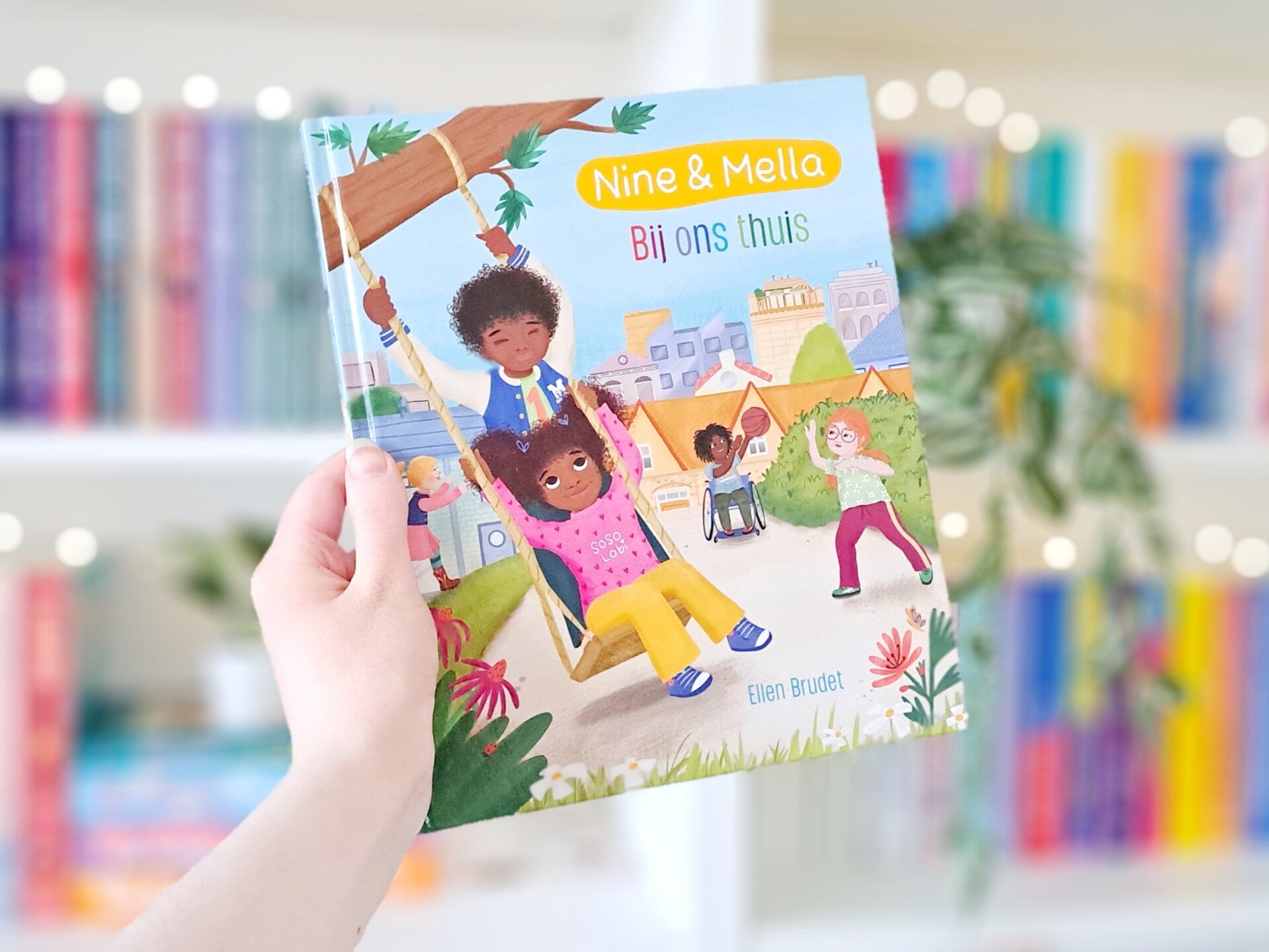 Een witte hand houdt 'Bij ons thuis' van Ellen Brudet en Maruga Koops omhoog voor een kleurrijke wazige boekenkast met lichtjes. Foto bij Own-voices recensie door turnthepages.nl Rolstoelgebruiker en disabled bookstagrammer en blogger.