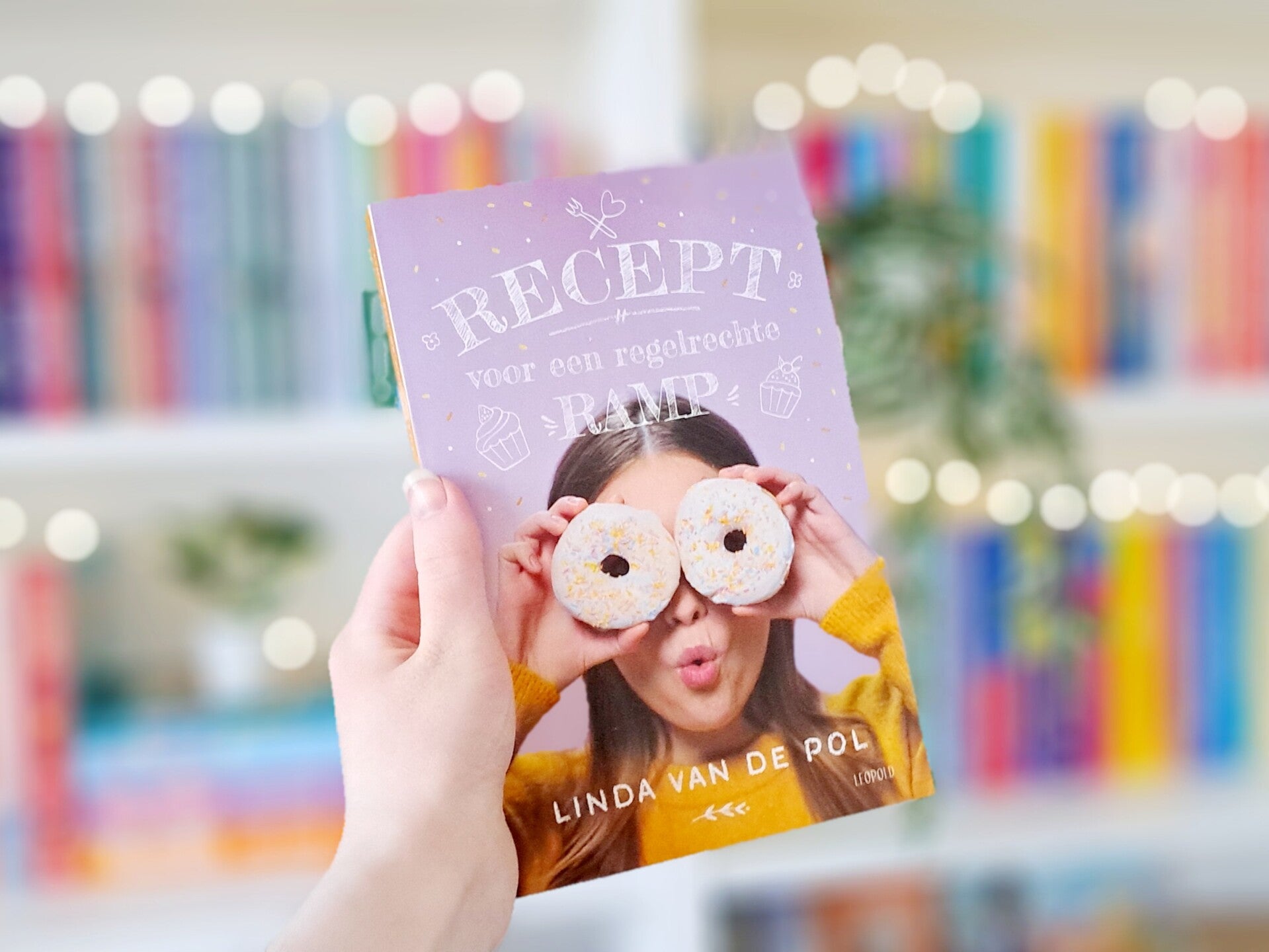 Een witte hand houdt 'Recept voor een regelrechte ramp' van Linda van de Pol omhoog voor een kleurrijke wazige boekenkast met lichjtes. Foto bij recensie door turnthepages.nl Rolstoelgebruiker en disabled bookstagrammer en blogger met ADHD.