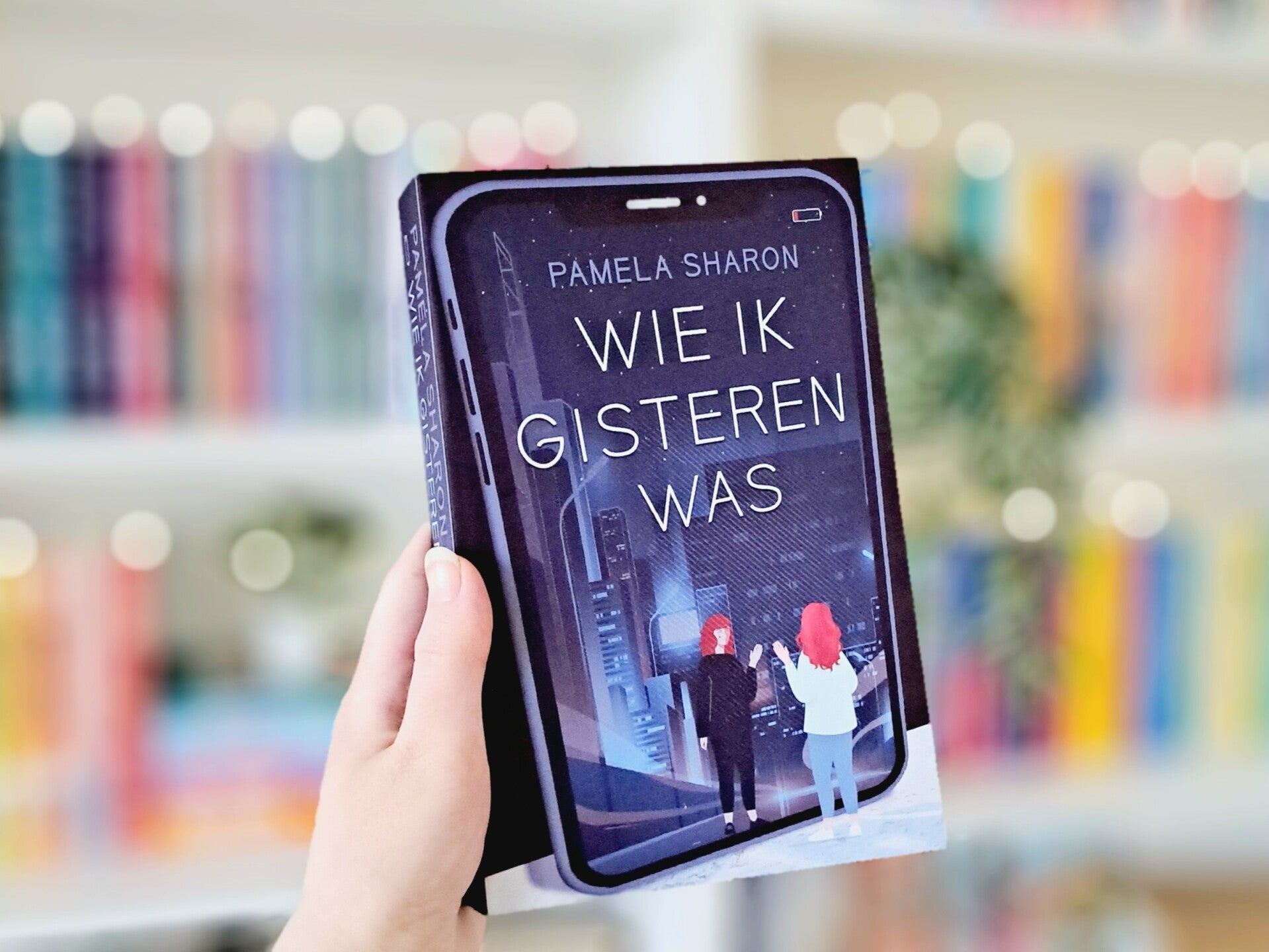 Een witte hand houdt 'Wie ik gisteren was'van Pamela Sharon omhoog voor een kleurrijke wazige boekenkast met lichtjes. Foto bij recensie door turnthepages.nl Rolstoelgebruiker en disabled bookstagrammer en blogger.