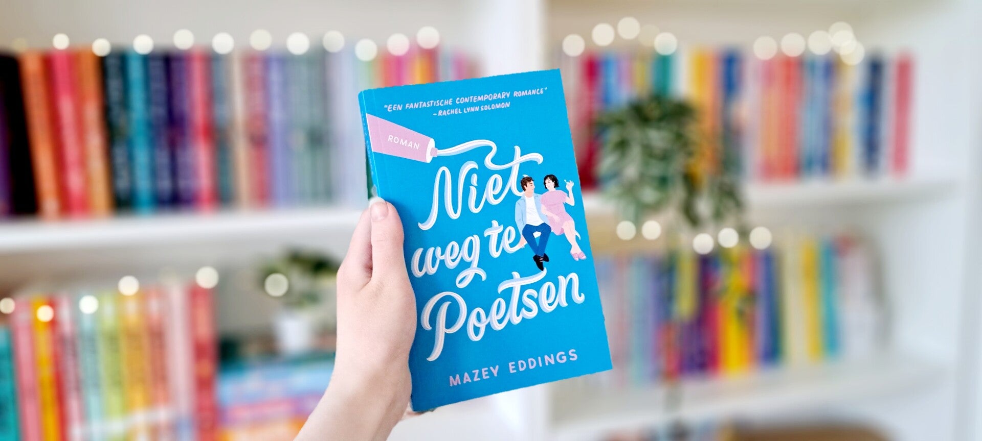 Een witte hand houdt 'Niet weg te poetsen' van Mazey Eddings omhoog voor een kleurrijke wazige boekenkast met lichtjes. Foto bij recensie door turnthepages.nl Rolstoelgebruiker, disabled bookstagrammer en queer blogger.