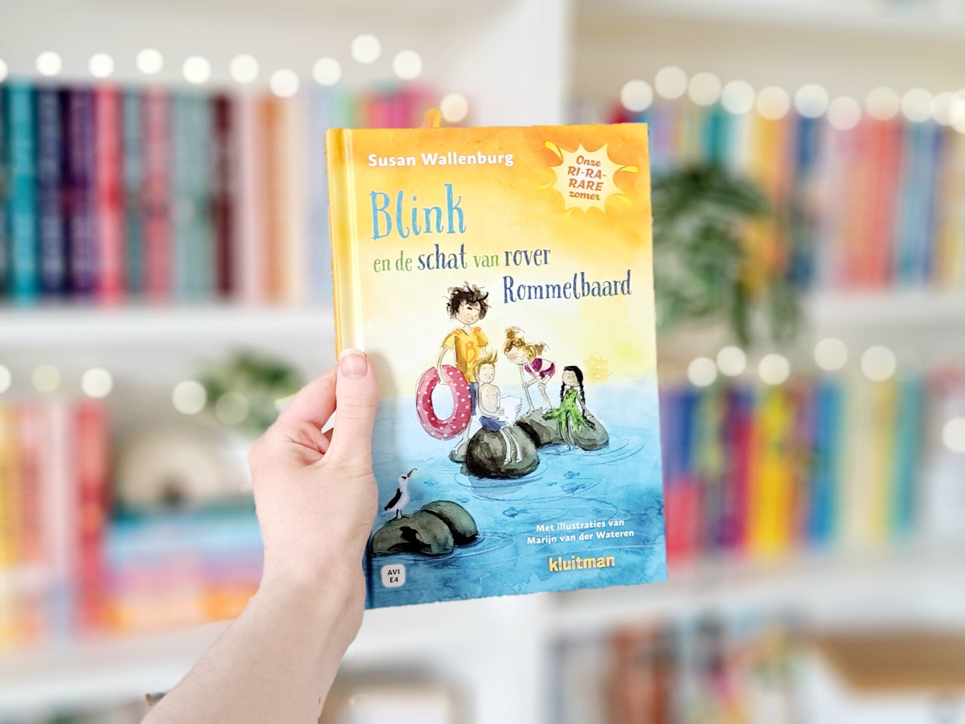 Een witte hand houdt 'Blink en de schat van rover Rommelbaard', deel 1 in de serie Onze Ri-ra-rare zomer van Susan Wallenburg. geïllustreerd door Marijn van der Wateren omhoog voor een kleurrijke wazige boekenkast met lichtjes. Foto bij own-voices recensi