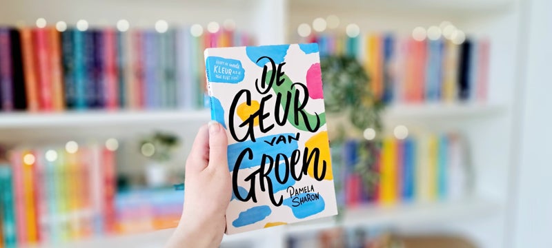Een witte hand houdt 'De geur van groen' van pamela Sharon omhoog voor een kleurrijke wazige boekenkast met kichtjes. Foto bij recensie door turnthepages.nl Rolstoelgebruiker en disabled bookstagrammer en blogger.