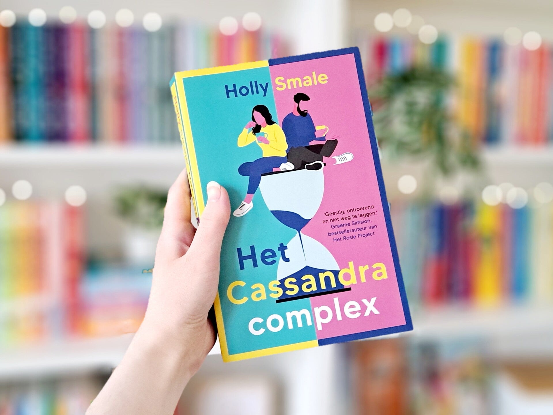Een witte hand houdt 'Het Cassandra complex' van Holly Smale omhoog voor een kleurrijke wazige boekenkast met lichtjes. Foto bij recensie door turnthepages.nl Rolstoelgebruiker en disabled bookstagrammer en blogger.