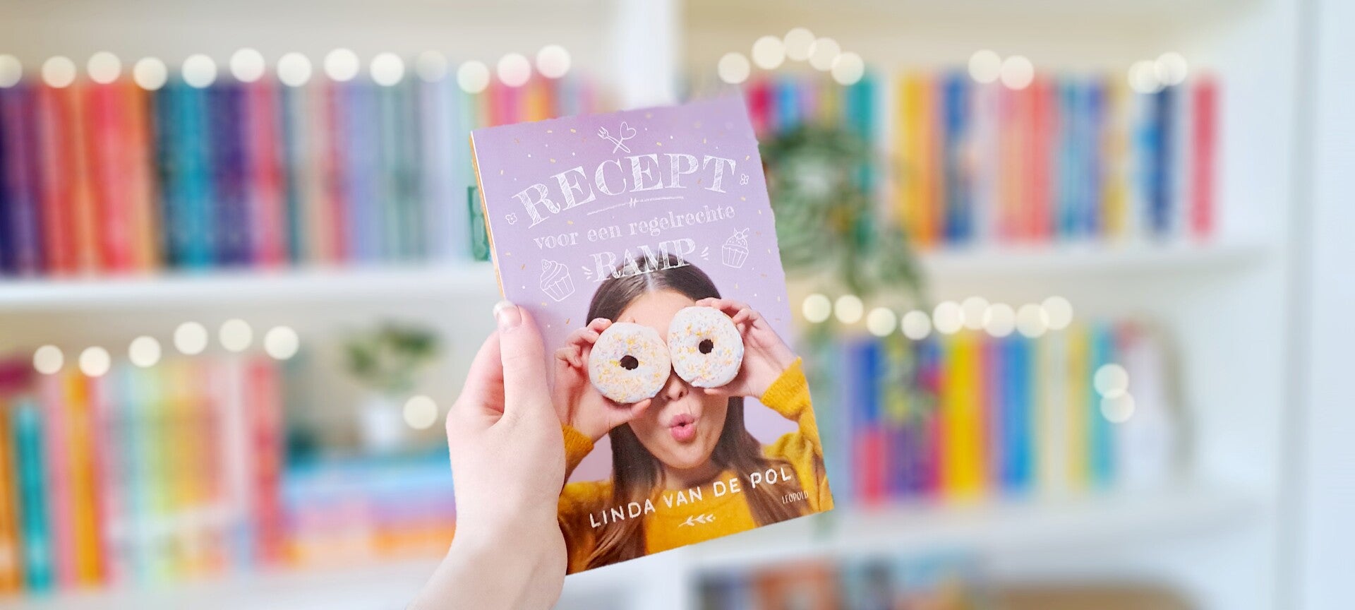 Een witte hand houdt 'Recept voor een regelrechte ramp' van Linda van de Pol omhoog voor een kleurrijke wazige boekenkast met lichtjes. Foto bij own-voices recensie door turnthepages.nl Rolstoelgebruiker en disabled bookstagrammer en blogger met ADHD.