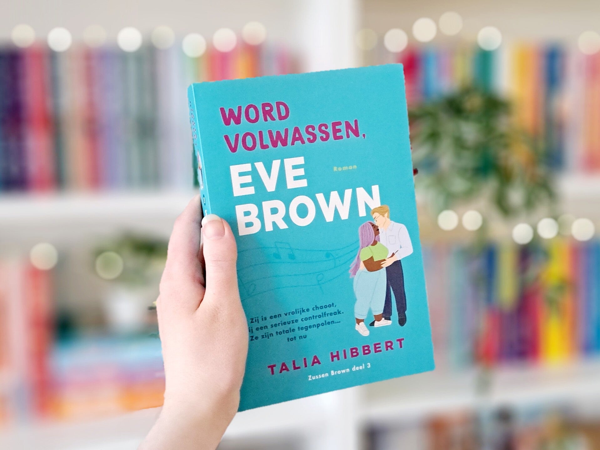 Een witte hand houdt 'Word volwassen, Eve Brown' deel drie in the sisters brown trilogie van Talia Hibbert omhoog voor een kleurrijke wazige boekenkast met lichtjes. Foto bij recensie door turnthepages.nl Rolstoelgebruiker en disabled bookstagrammer en bl