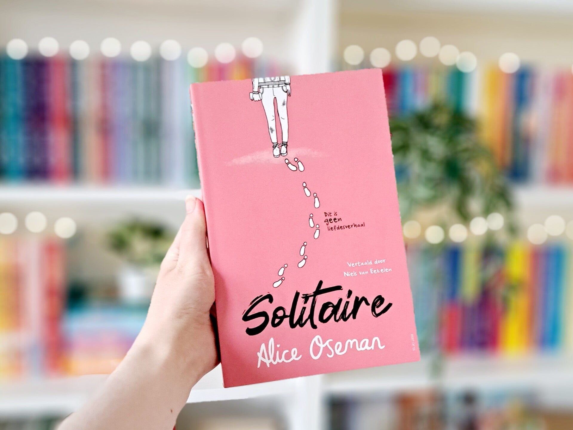 Een witte hand houdt 'Solitaire' van Alice Oseman omhoog voor een ekleurrijke wazige boekenkast met lichtjes. Foto bij recensie door turnthepages.nl Disabled bookstagrammer en queer blogger.