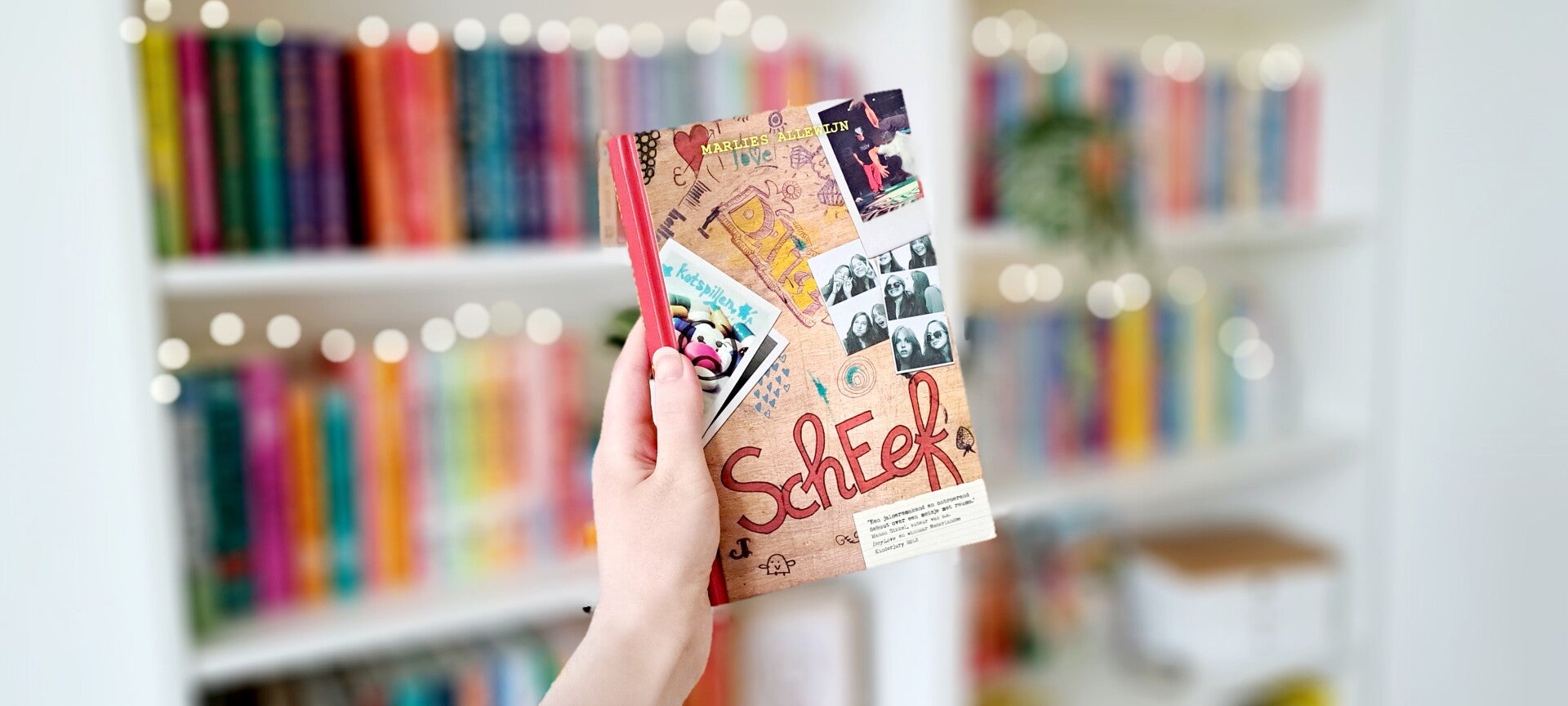 Een witte hand houdt 'SchEef' van Marlies Allewijn omhoog voore en kleurrijke wazige boekenkast met lihcjtes. Foto bij recensie door tutnrhtpegae.nl Rolstoelgebruiker en disabled bookstagrammer en blogger.