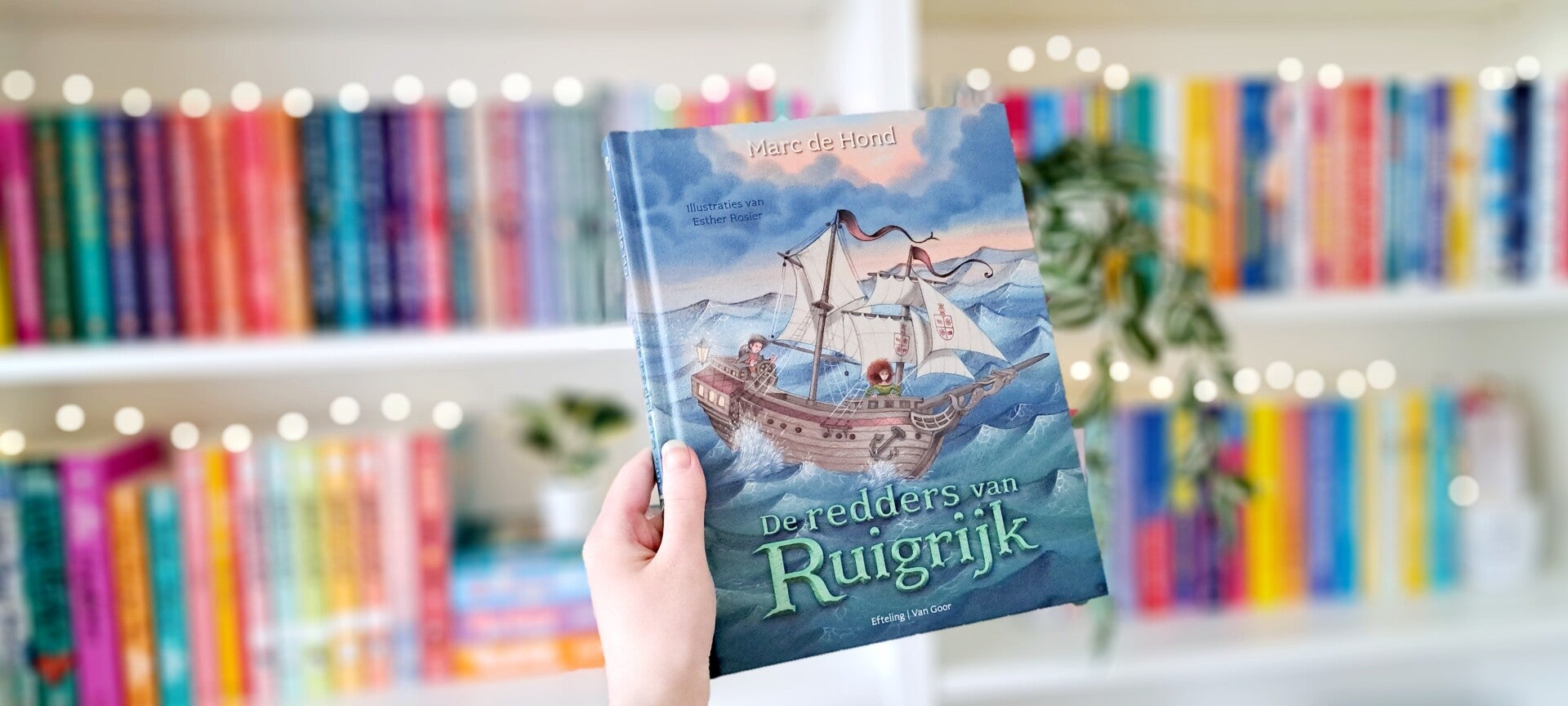 Een witte hand houdt 'De redders van Ruigrijk' van Marc de Hond omhoog voor een kleurrijke wazige boekenkast met lichtjes. Foto bij own-voices recensie door turnthepages.nl Rolstoelgebruiker en disabled bookstagrammer en blogger.