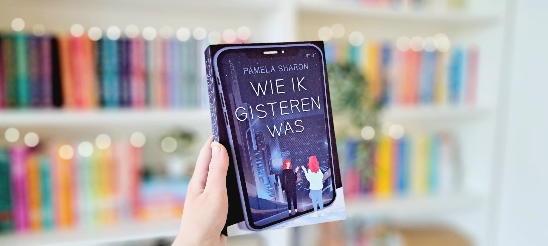 Een witte hand houdt 'Wie ik gisteren was'van Pamela Sharon omhoog voor een kleurrijke wazige boekenkast met lichtjes. Foto bij recensie door turnthepages.nl Rolstoelgebruiker en disabled bookstagrammer en blogger.