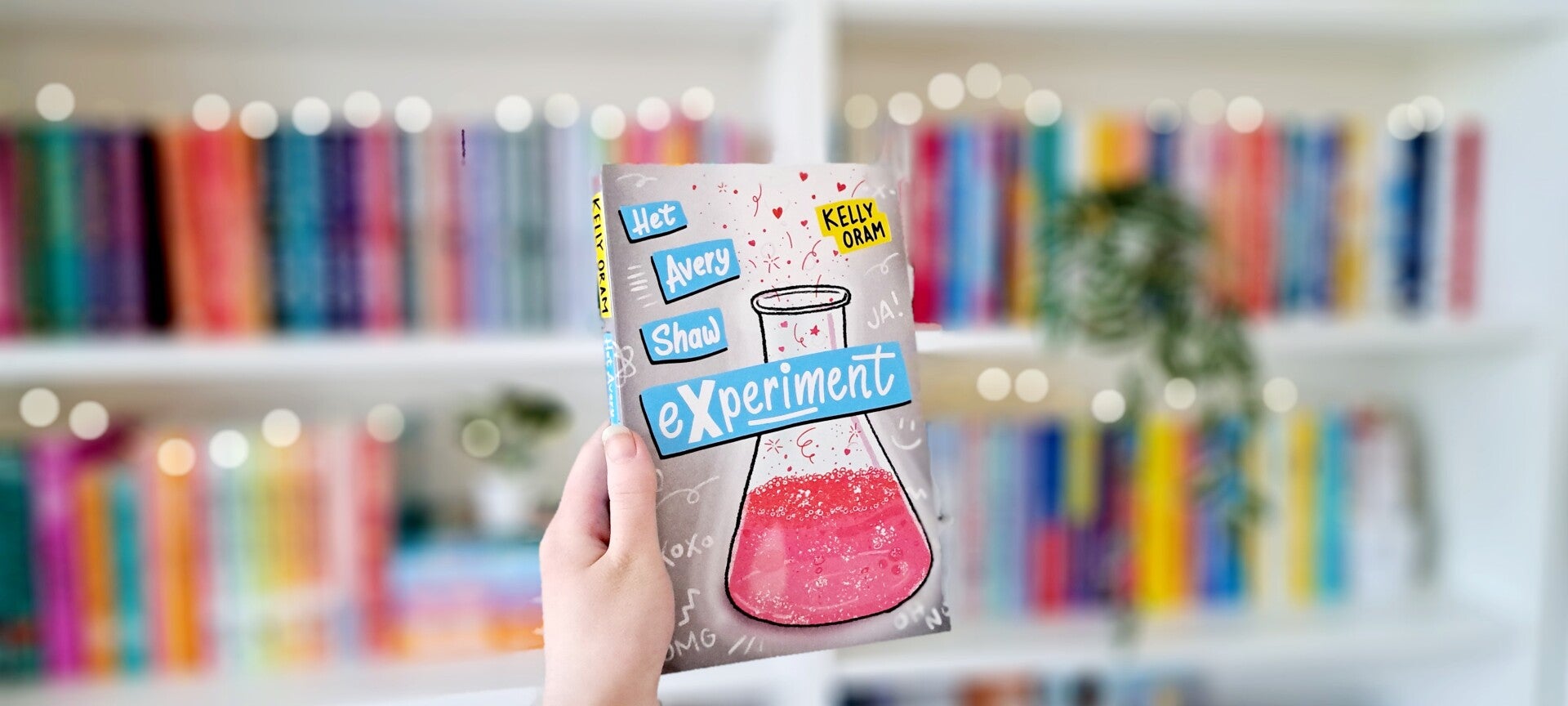 Een witte hand houdt 'Het Avery Shaw experiment' van Kelly Oram omhoog voor een kleurrijke wazige boekenkast met lichtjes. Foto bij recensie door turnthepages.nl Rolstoelgebruiker, disabled bookstagrammer en queer blogger.