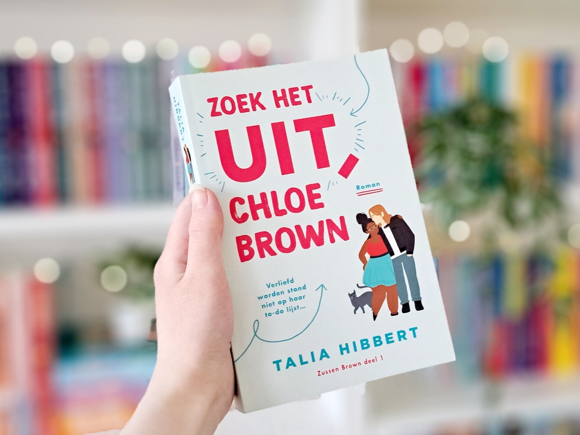 Een witte hand houd 'Zoek het uit, Chloe Brown' van Talia Hibbert omhoog voor een kleurrijke wazige boekenkast met lichtjes. Foto bijown-voices  recensie door turnthepages.nl Rolstoelgebruiker en disabled bookstagrammer en blogger.