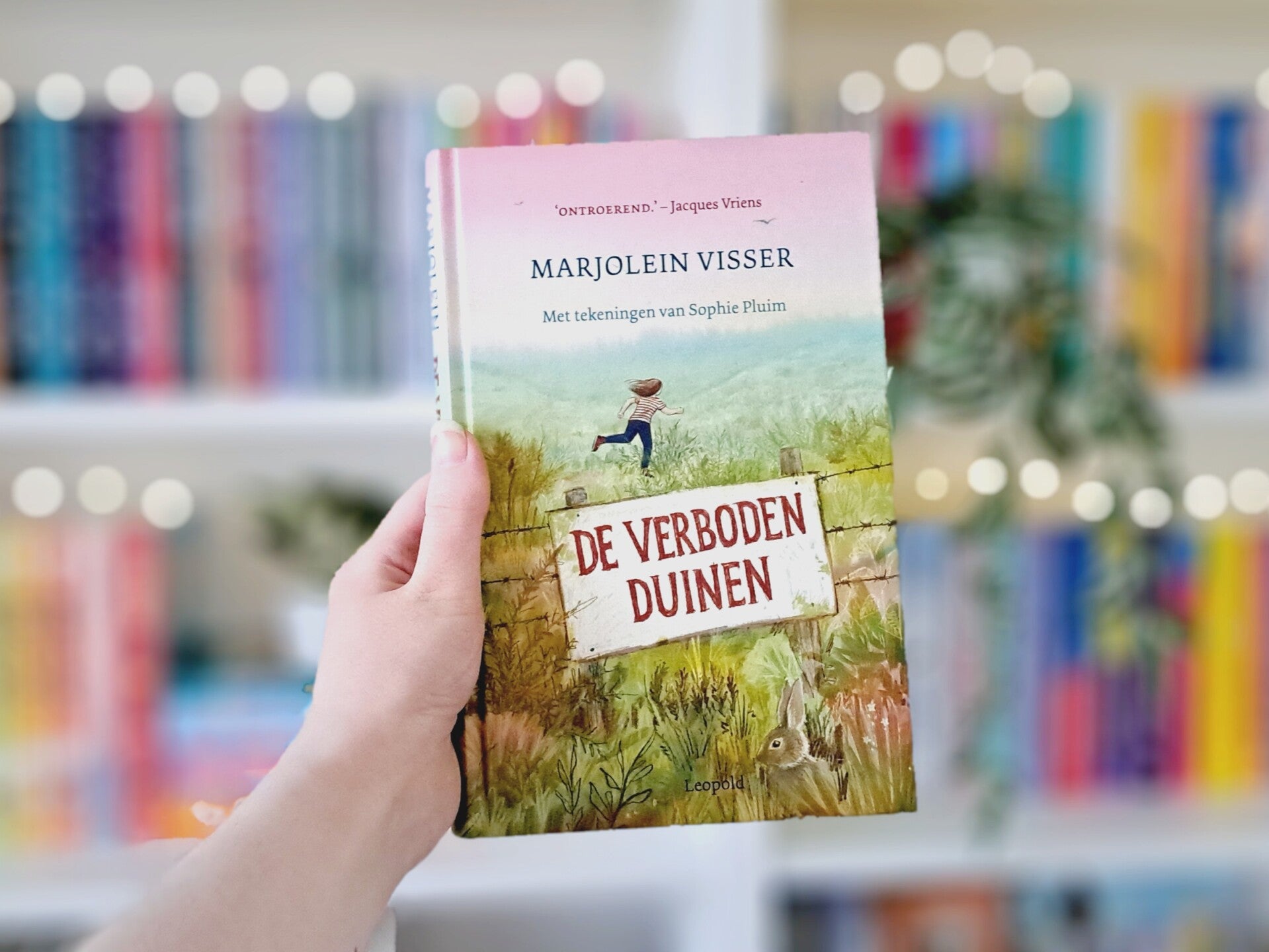 Een witte hand houdt 'De verboden duinen' van Marjolein Visser omhoog voor een kleurrijke wazige boekenkast met lihcjtes. Foto bij recensie door turnthepages.nl Rolstoelgebruiker en disabled bookstagrammer en blogger.