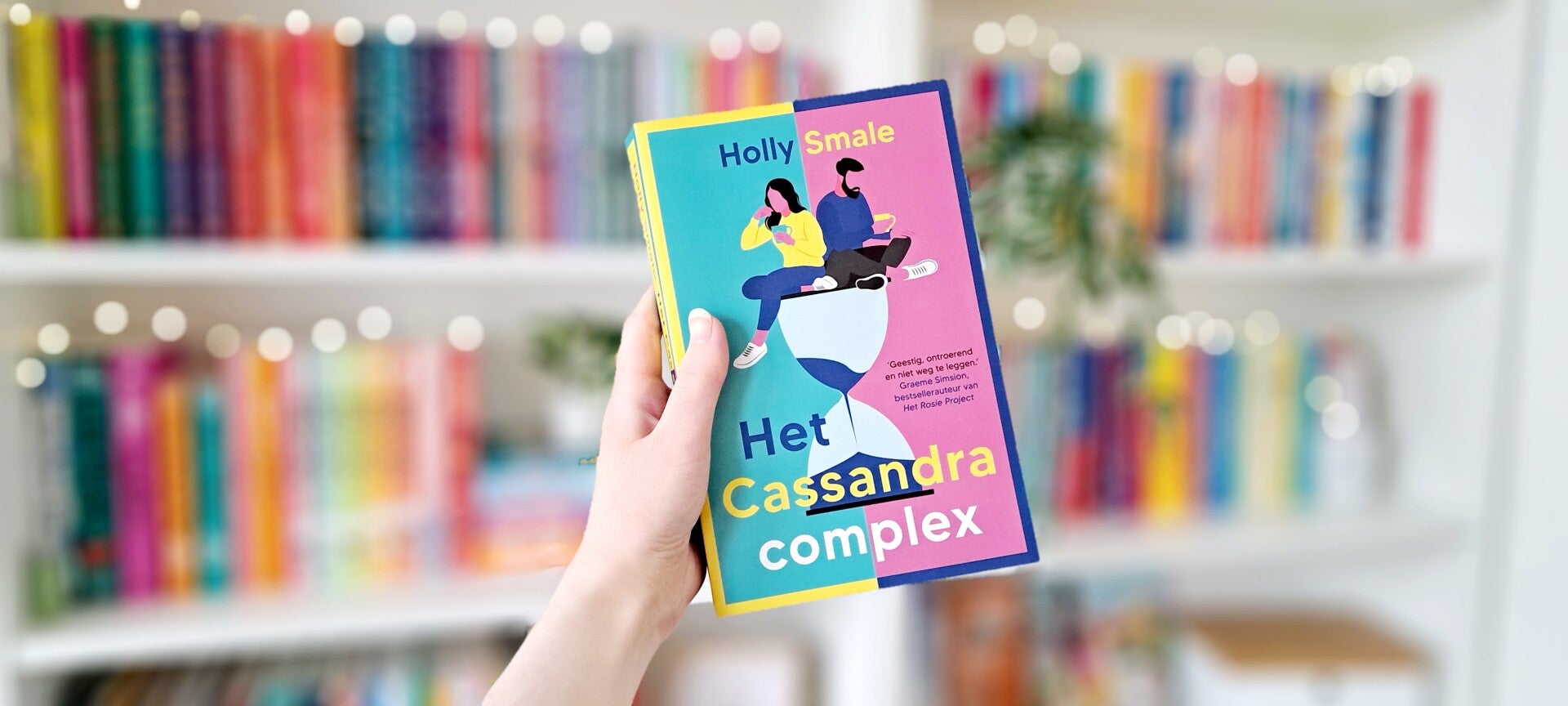 Een witte hand houdt 'Het Cassandra complex' van Holly Smale omhoog voor een kleurrijke wazige boekenkast met lichtjes. Foto bij recensie door turnthepages.nl Rolstoelgebruiker en disabled bookstagrammer en blogger.