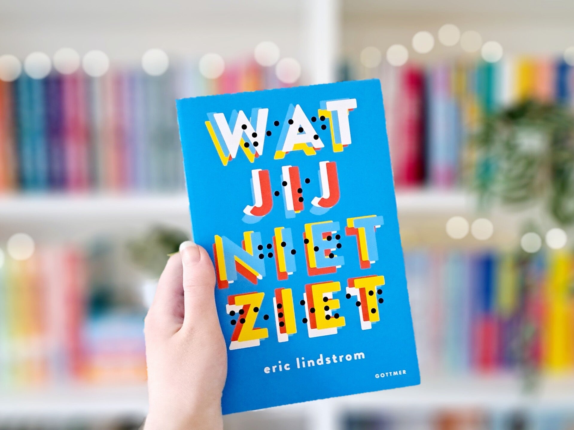 Een witte hand houdt 'Wat jij niet ziet' van Eric Lindström omhoog voor een kleurrijke wazige boekenkast met lichtjtes. Foto bij recensie door turnthepages.nl Rolstoelgebruiker en disabled bookstagrammer en blogger.
