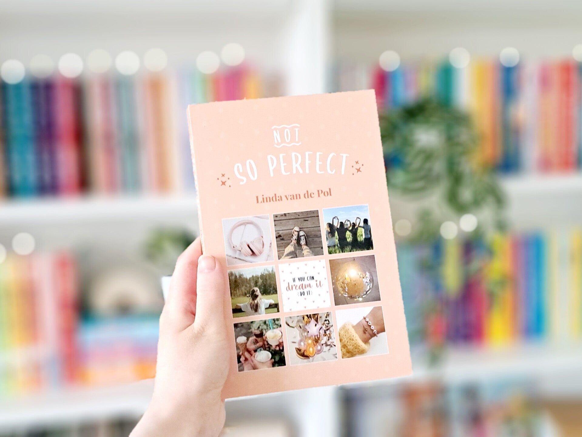 Een witte hand houdt '(Not) so perfect' van Linda van de Pol omhoog voor een kleurrijke wazige boekenkast met lichtjes. Foto bij recensie door turnthepages.nl Disabled bookstagrammer en boekblogger.