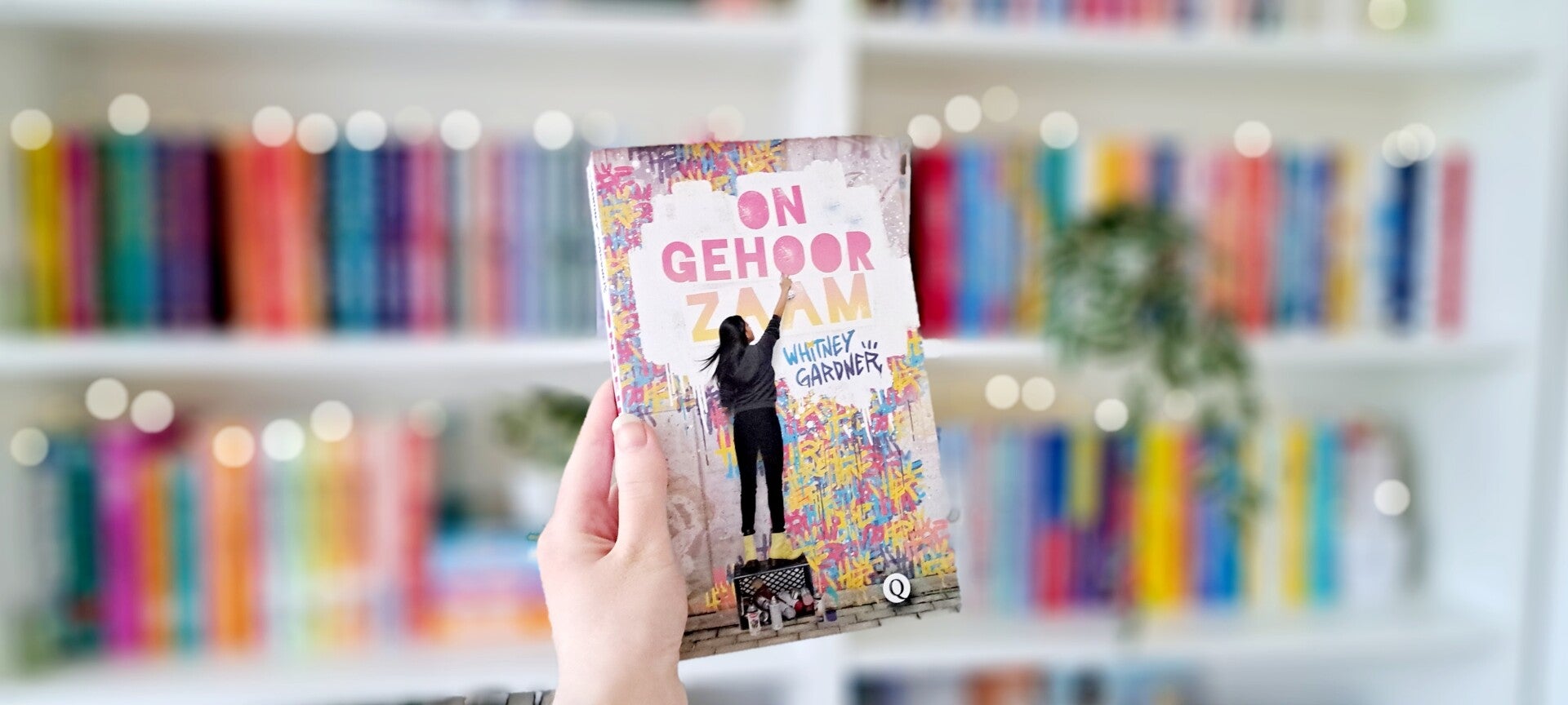 Een witte hand houdt 'Ongehoorzaam' van Whitney Gardner omhoog voor een kleurrijke wazige boekenkast met lichtjes. Foto bij recensie door turnthepages.nl Rolstoelgebruiker en disabled bookstagrammer en blogger.