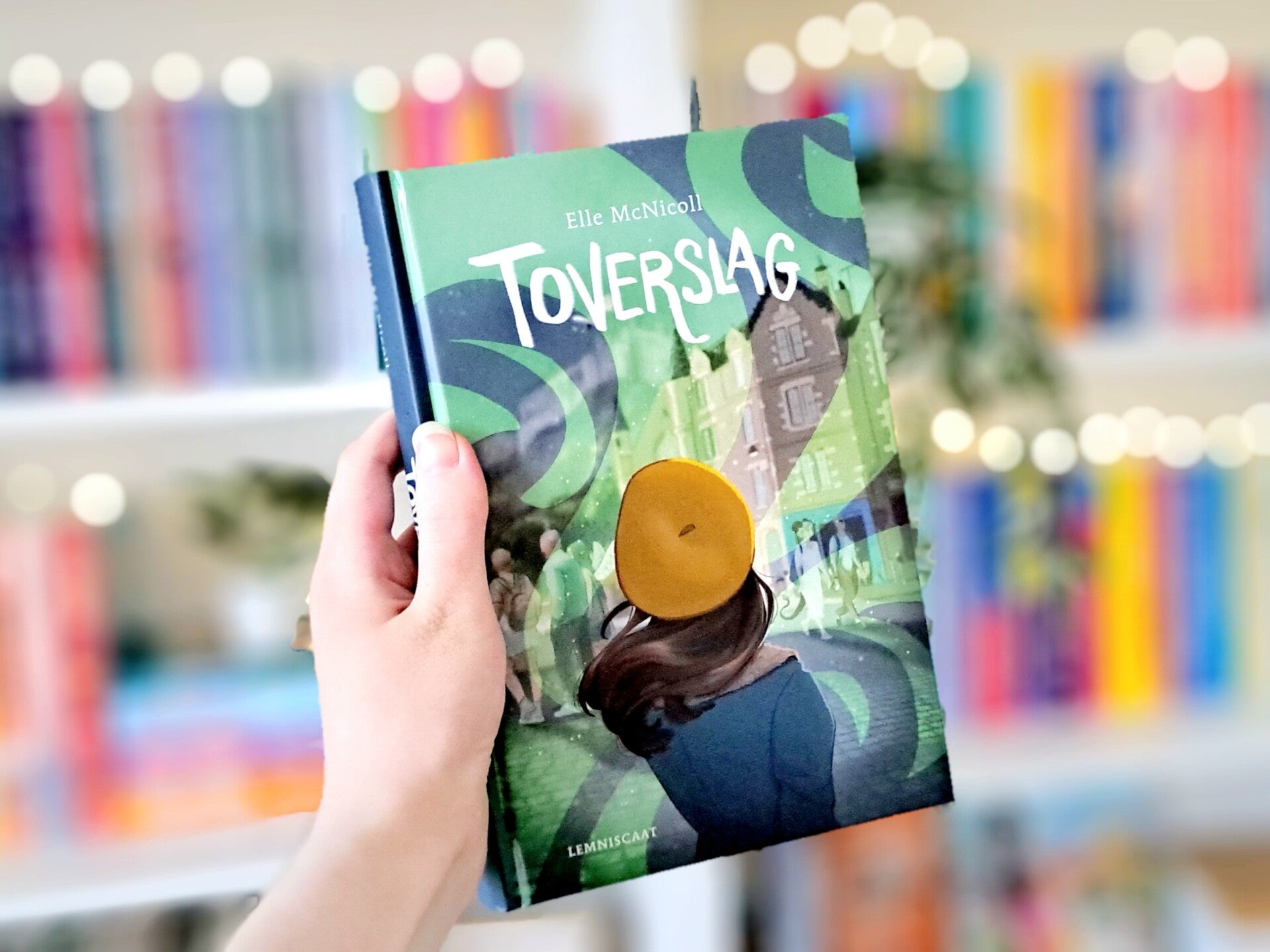 Een witte hand houdt 'Toverslag' van Elle McNicoll omhoog voor een kleurrijke wazige boekenkast met lichtjes. Foto bij recensie door turnthepages.nl Rolstoelgebruiker en neurodivergente disabled bookstagrammer en blogger.
