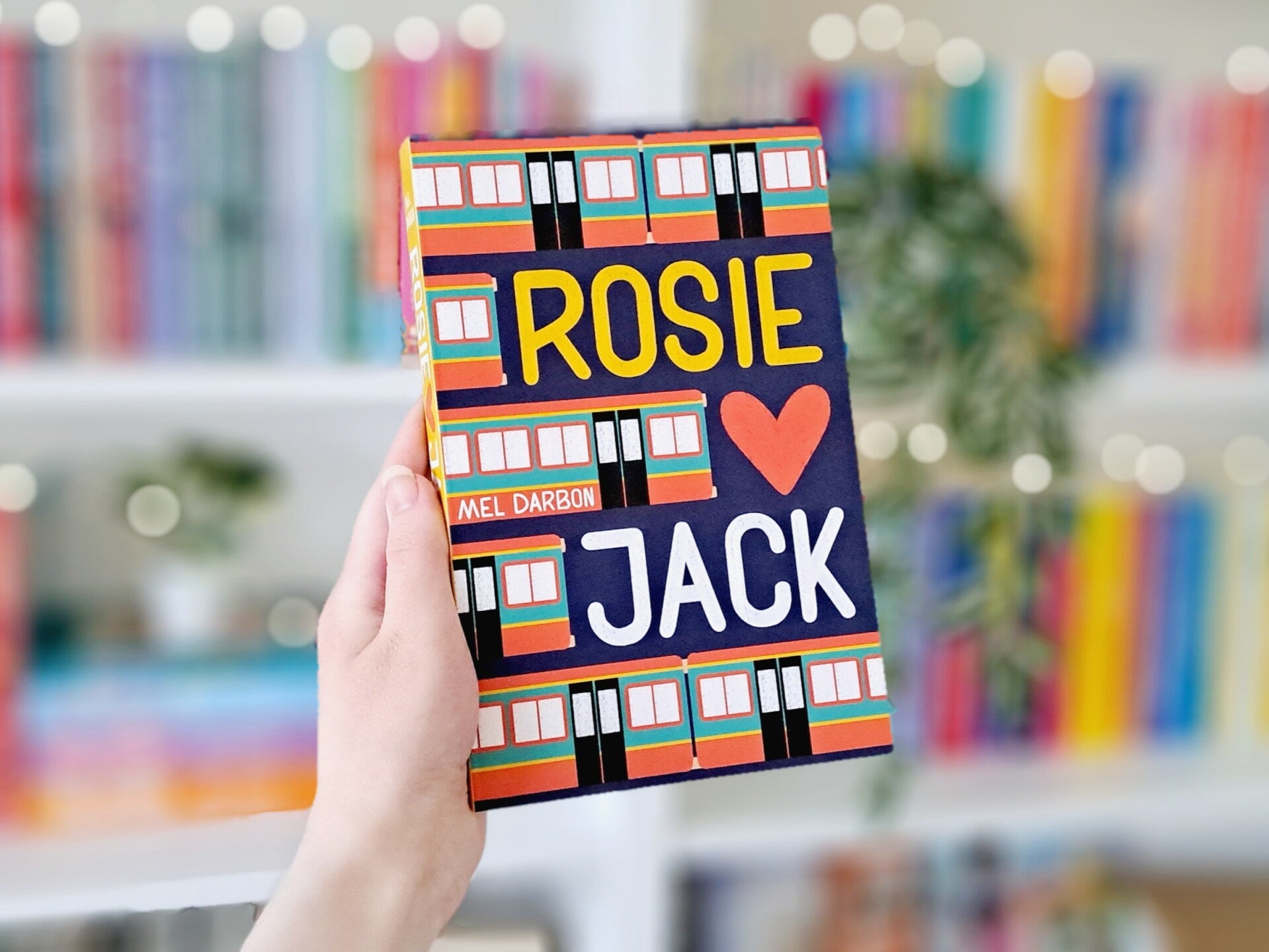 Een witte hand houdt 'Rosie hartje Jack' van mel Darbon omhoog voor een kleurrijke wazige boekenkast met lichtjes. Foto bij recensie door turnthepages.nl Rolstoelgebruiker en disabled bookstagrammer en blogger.