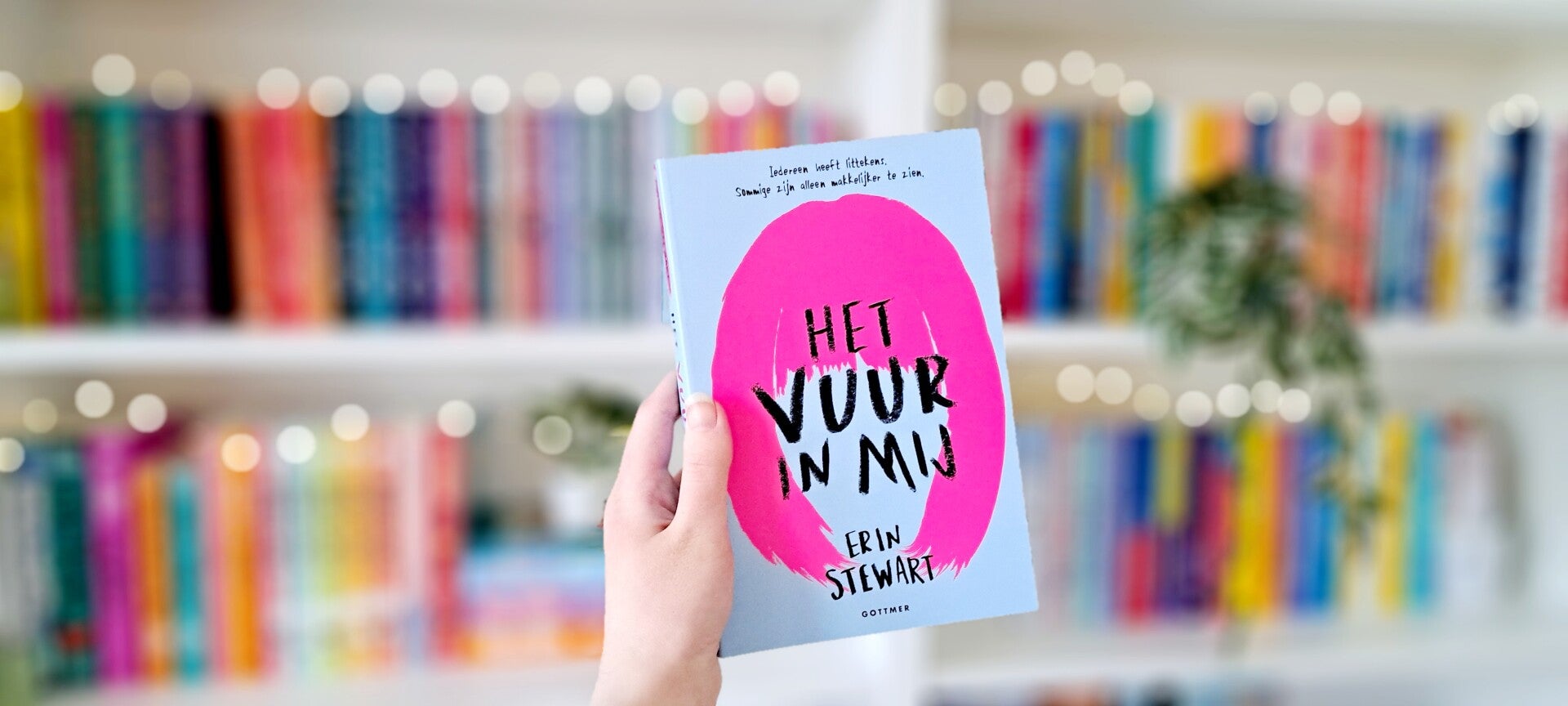 Een witte hand houdt 'Het vuur in mij' geschreven door Erin Stewart, omhoog voor een kleurrijke wazige boekenkast met lichtjes. Foto bij recensie door turnthepages.nl Rolstoelgebruiker en disabled bookstagrammer en blogger.