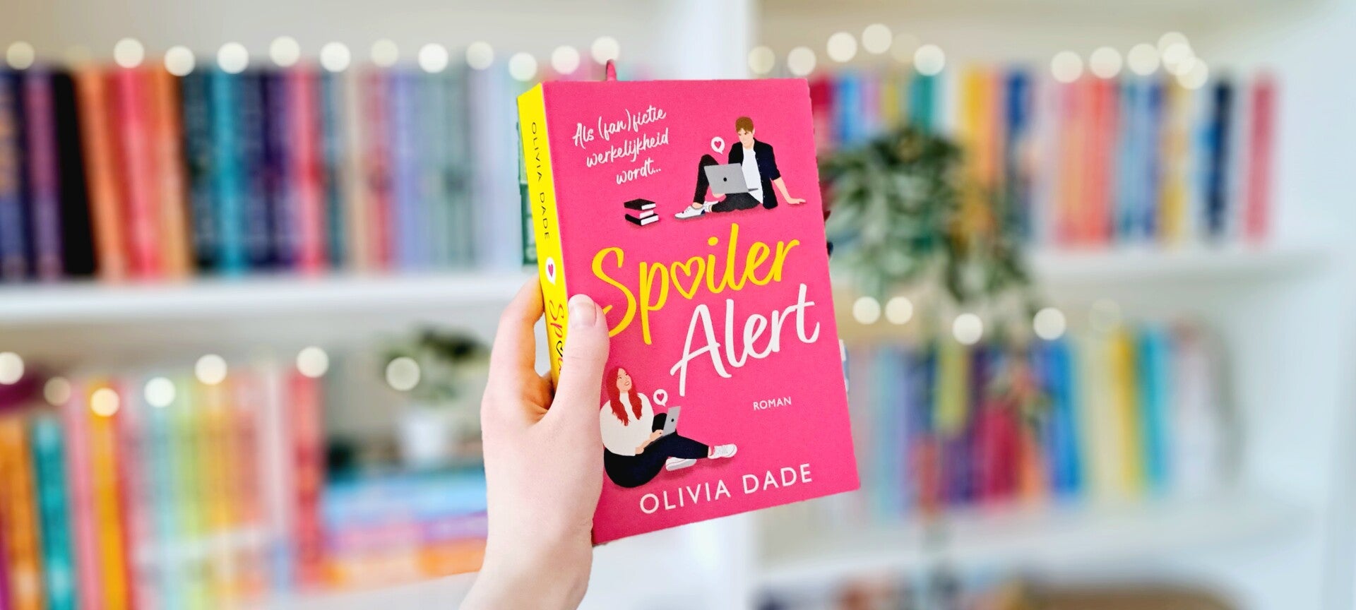 Een witte hand houdt 'Spoiler alert' van Olivia Dade omhoog voor een kleurrijke wazige boekenkast met lihcjtes. Foto bij recensie door turnthepages.nl Rolstoelgebruiker en disabled bookstagrammer en blogger.