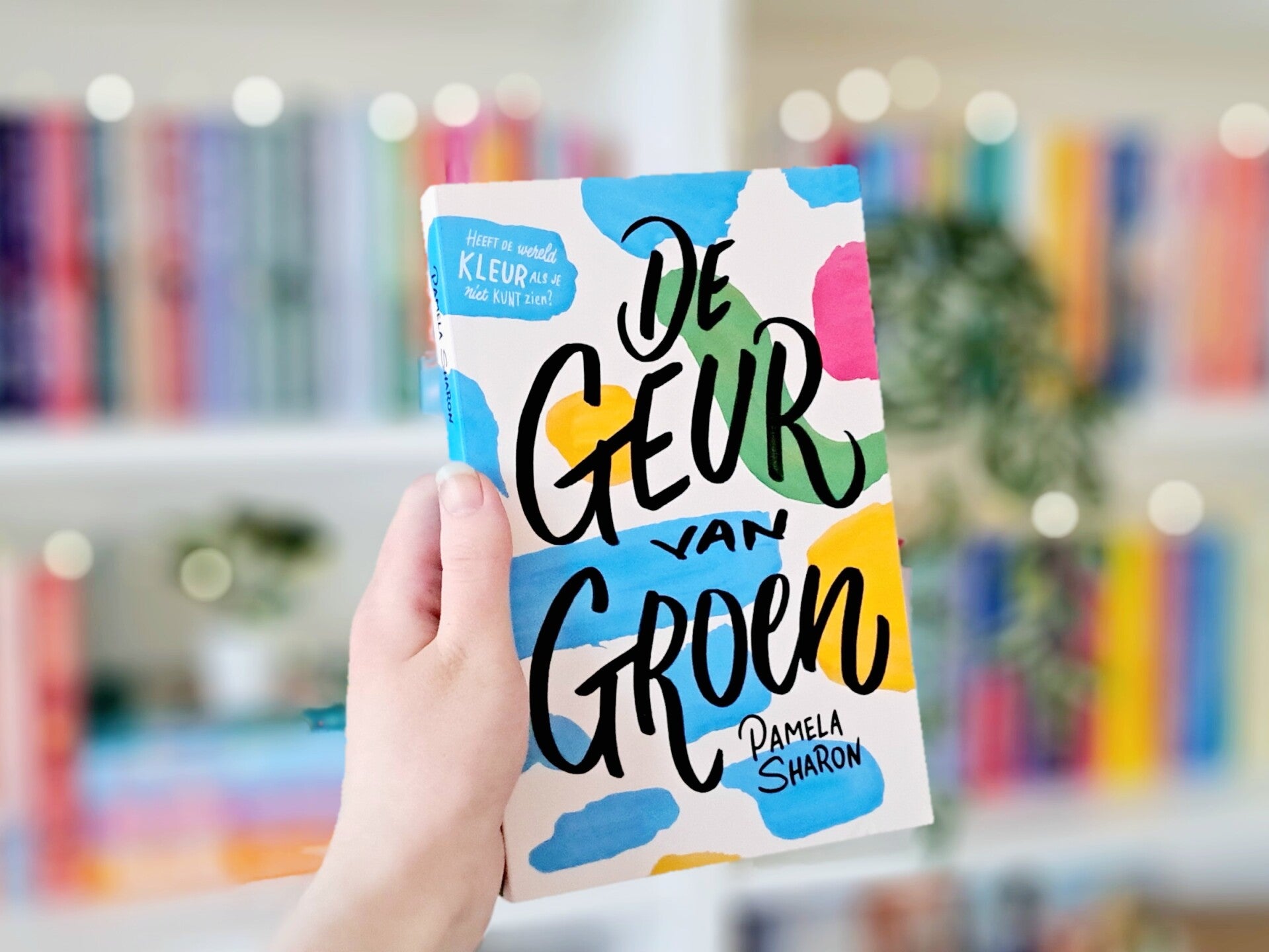Een witte hand houdt 'De geur van groen' van pamela Sharon omhoog voor een kleurrijke wazige boekenkast met kichtjes. Foto bij recensie door turnthepages.nl Rolstoelgebruiker en disabled bookstagrammer en blogger.