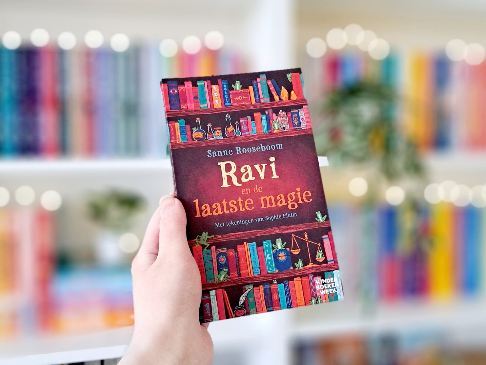 Een witte hand houdt 'Ravi en de laatste magie' van Sanne Rooseboom omhoog voor een kleurrijke wazige boekenkast met lichtjes. Foto bij recensie door turnthepages.nl Rolstoelgebruiker en disabled bookstagrammer en blogger.