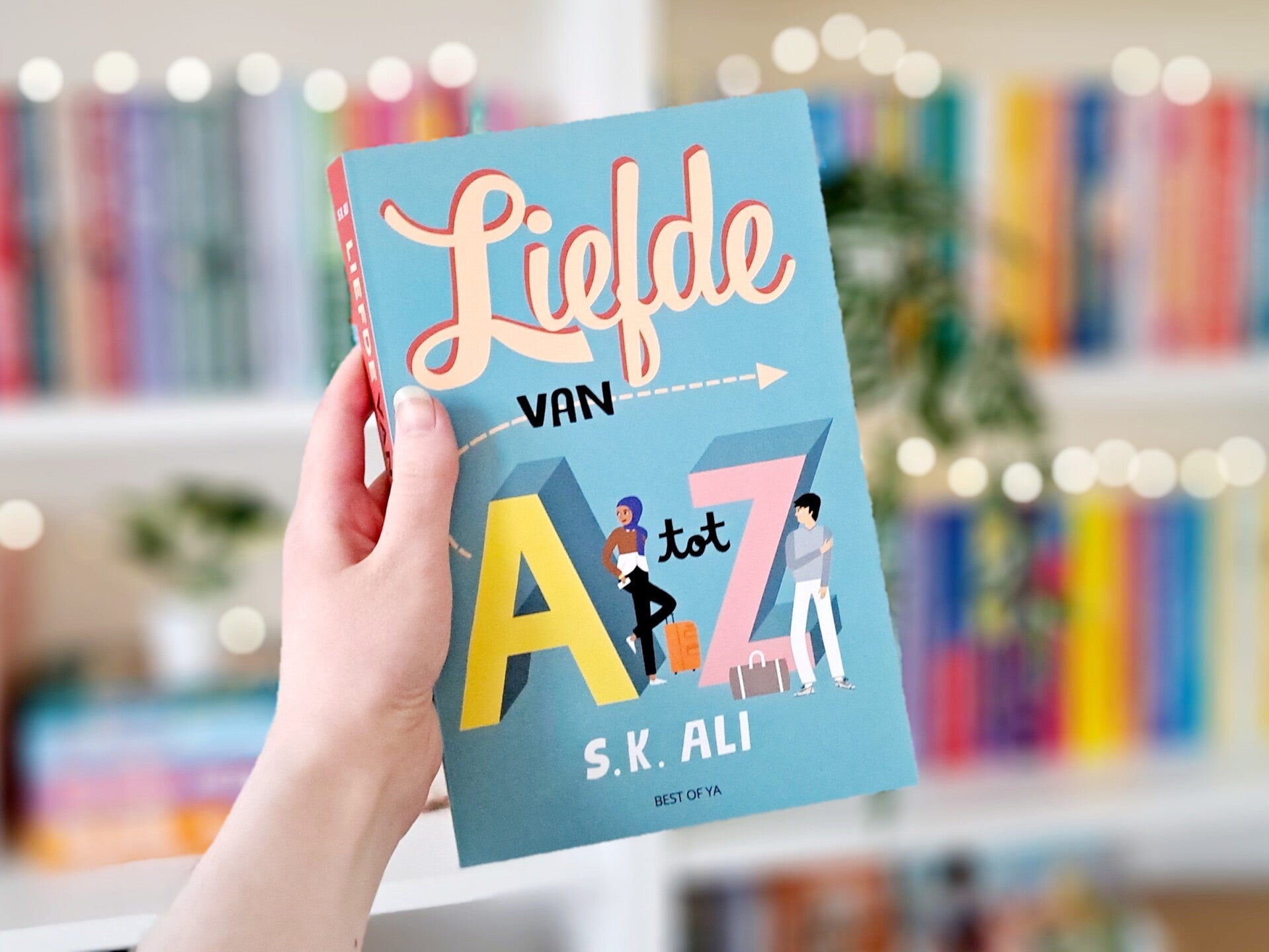 Een witte hand houdt 'Liefde van A tot Z' van S.K. Ali omhoog voor een kleurrijke wazige boekenkast met lichtjes. Foto bij recensie door turnthepages.nl Rolstoelgebruiker en disabled bookstagrammer en blogger.