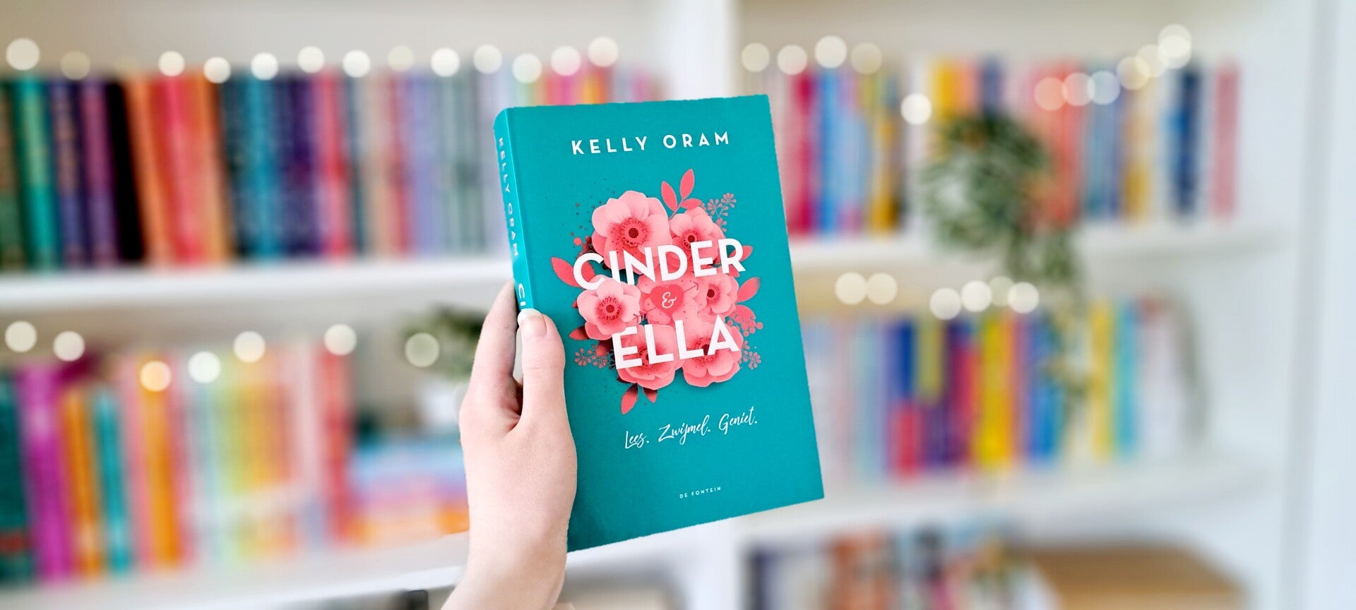 Een witte hand houdt 'Cinder & Ella' van Kelly Oram omhoog voor een kleurrijke wazige boekenkast met lichtjes. Foto bij personage quiz door turnthepages.nl Disabled bookstagrammer en boekblogger.