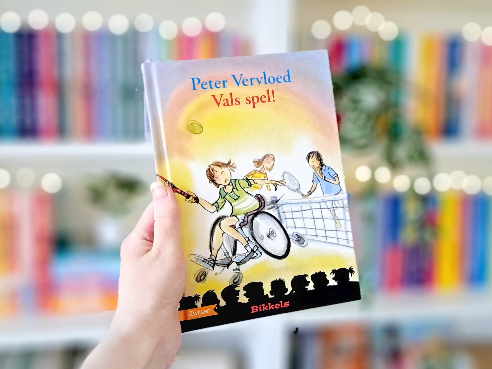 Een witte hand houdt 'Vals spel!' van Peter Vervloed omhoog voor een kleurrijke wazige boekenkast met lichtjes. Foto bij recensie door turnthepages.nl Rolstoelgebruiker en disabled bookstagrammer en blogger.