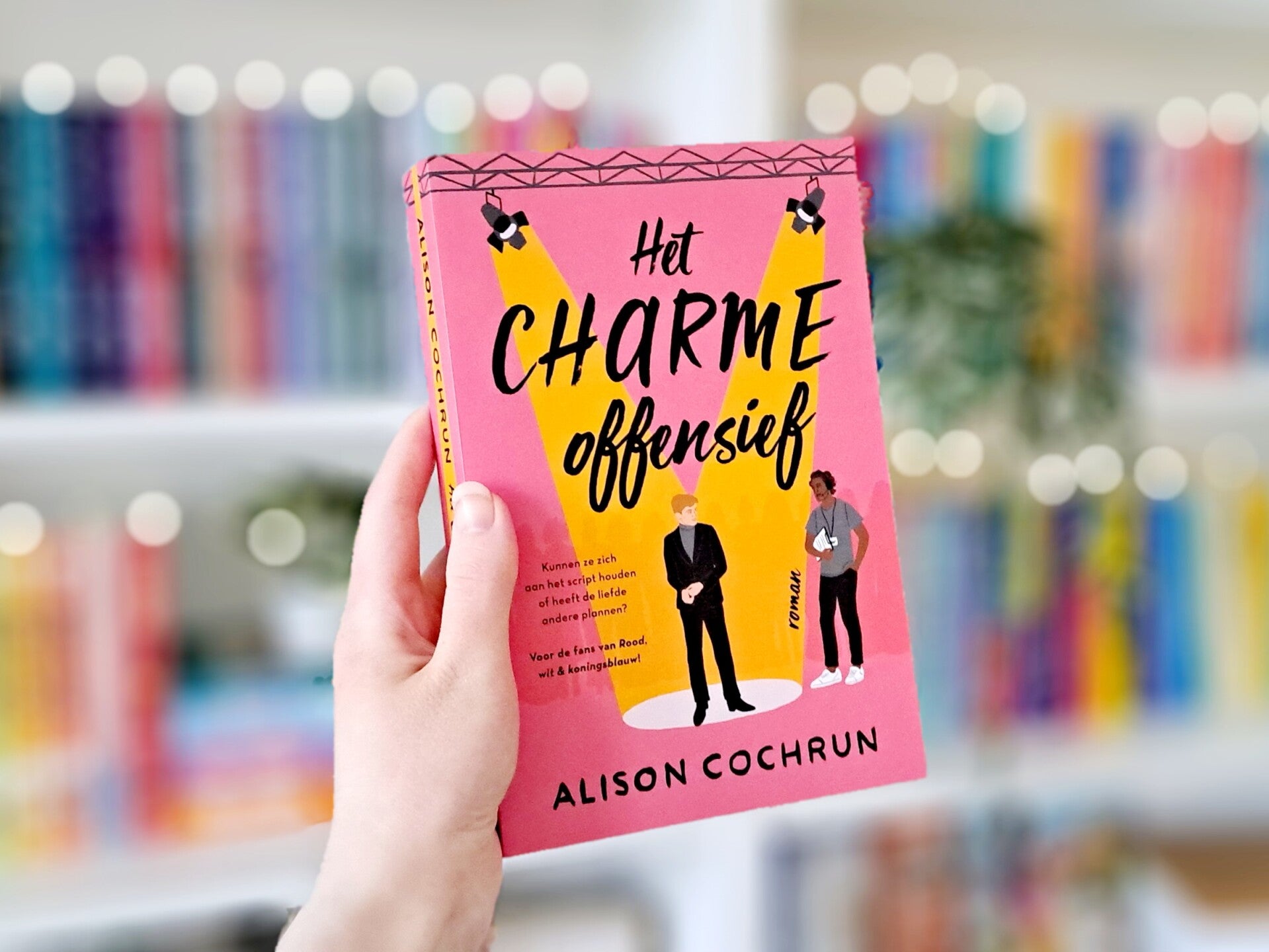 Een witte hand houdt 'Het charmeoffensief' van Alison Cochrun omhoog voor een kleurrijke wazige boekenkast met lichtjes. Foto bij recensie door turnthepages.nl Rolstoelgebruiker en disabled bookstagrammer en blogger met ADHD.