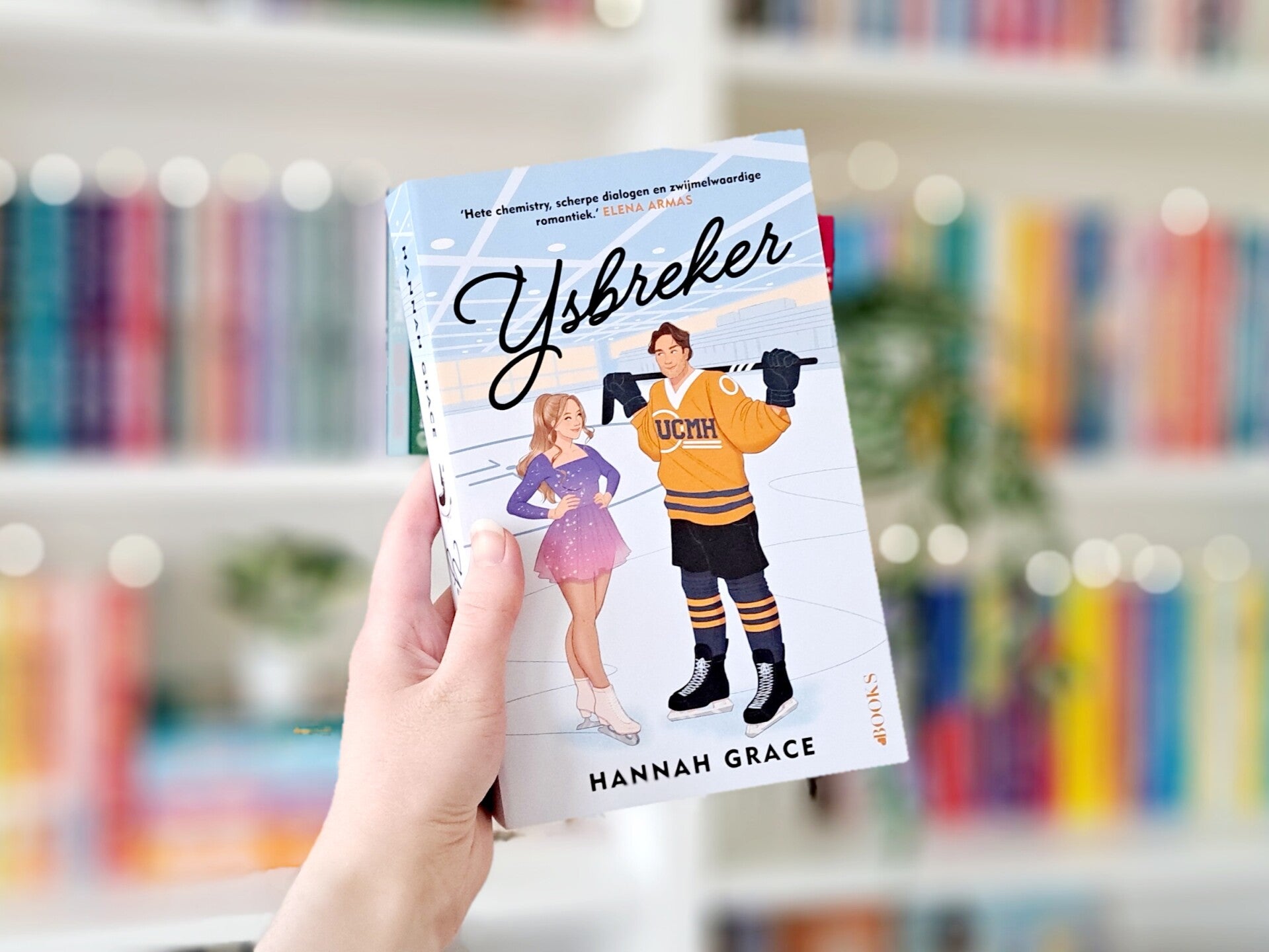 Een witte hand houdt 'IJsbreker' van Hannah Grace voor een kleurrijke wazige boekenkast met lichtjes. Foto bij recensie van turnthepages.nl Rolstoelgebruiker en disabled bookstagrammer en blogger.