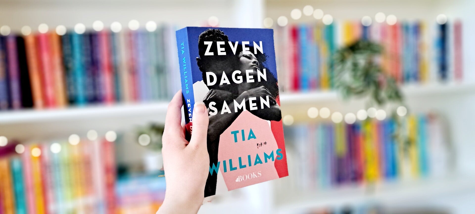 Een witte hand houdt 'Zeven dagen samen' van Tia WIlliams omhoog voor een kleurrijke wazige boekenkast met lichtjes. Recensie door turnthepages.nl Rolstoelgebruiker en disabled bookstagrammer en blogger.