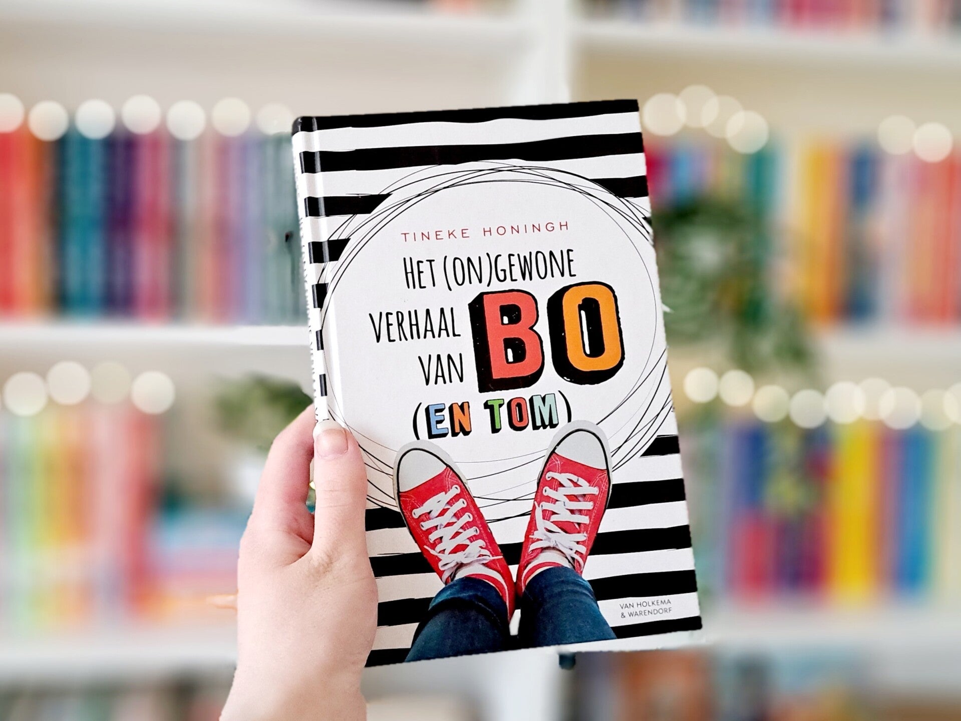 Een witte hand houdt 'het (on)gewone verhaal van Bo (en Tom) van Tineke Honingh omhoog tegen een wazige boekenkast met lichtjes. Foto bij recensie van turnthepages.nl Rolstoelgebruiker en disabled bookstagrammer en blogger.