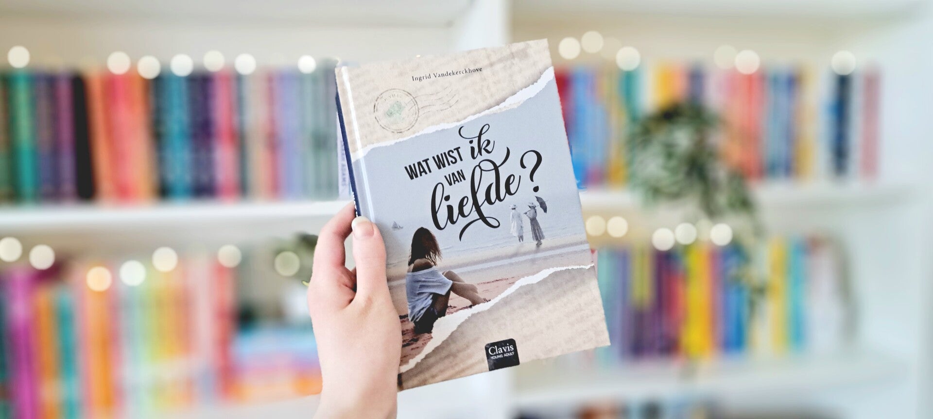 Een witte hand houdt 'Wat wist ik van liefde?' van Ingrid Vandekerckhove omhoog voor een kleurrijke wazige boekenkast met lichtjes. Foto bij recensie door turnthepages.nl Rolstoelgebruiker, disabled bookstagrammer en queer blogger.