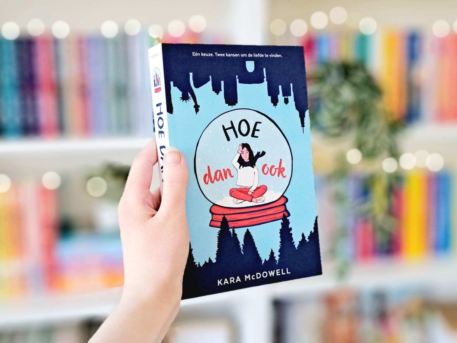 Een witte hand houdt 'Hoe dan ook' van Kara McDowell omhoog voor een kleurrijke wazige boekenkast met lichtjes. Foto bij recensie door turnthepages.nl Rolstoelgebruiker, disabled bookstagrammer en queer blogger.