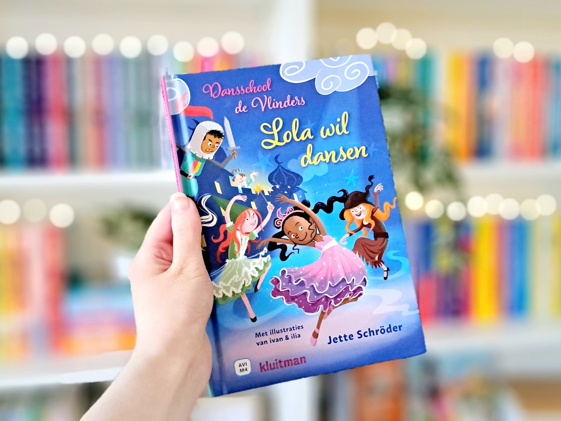 Een witte hand houdt 'Lola wil dansen' 'dansschool de vlinders deel 1'van Jette Schröder omhoog voor een kleurrijke wazige boekenkast met lichtjes. Foto bij recensie door turnthepages.nl Rolstoelgebruiker en disabled bookstagrammer en blogger.