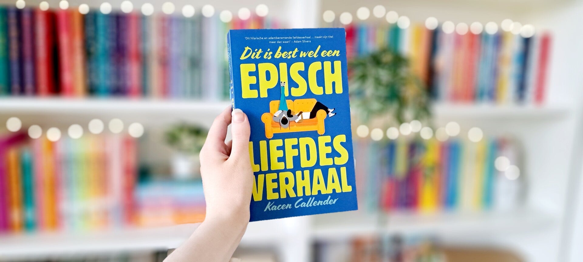 Een witte hand houdt 'Dit is best wel een episch liefdesverhaal' van Kacen Callender omhoog voor een kleurrijke wazige boekenkast met lichtjes. Foto bij recensie door turnthepages.nl Rolstoelgebruiker en disabled bookstagrammer en blogger.