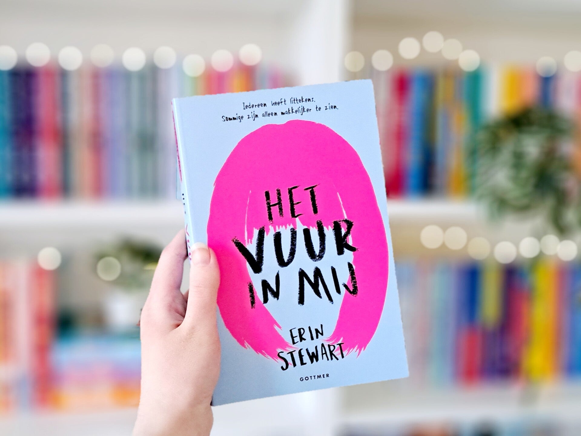 Een witte hand houdt 'Het vuur in mij' van Erin Stewart omhoog voor een kleurrijke wazige boekenkast met lichtjes. Foto bij recensie door turnthepages.nl Rolstoelgebruiker, disabled bookstagrammer en boekblogger.