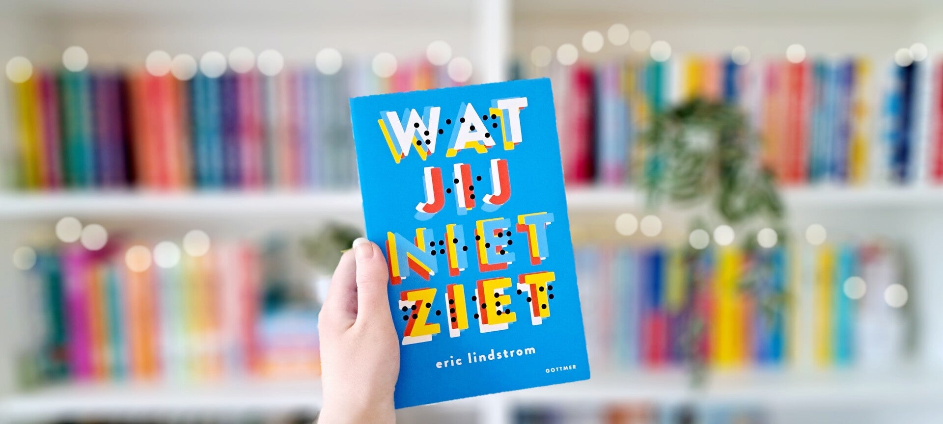 Een witte hand houdt 'Wat jij niet ziet' van Eric Lindström omhoog voor een kleurrijke wazige boekenkast met lichtjtes. Foto bij recensie door turnthepages.nl Rolstoelgebruiker en disabled bookstagrammer en blogger.