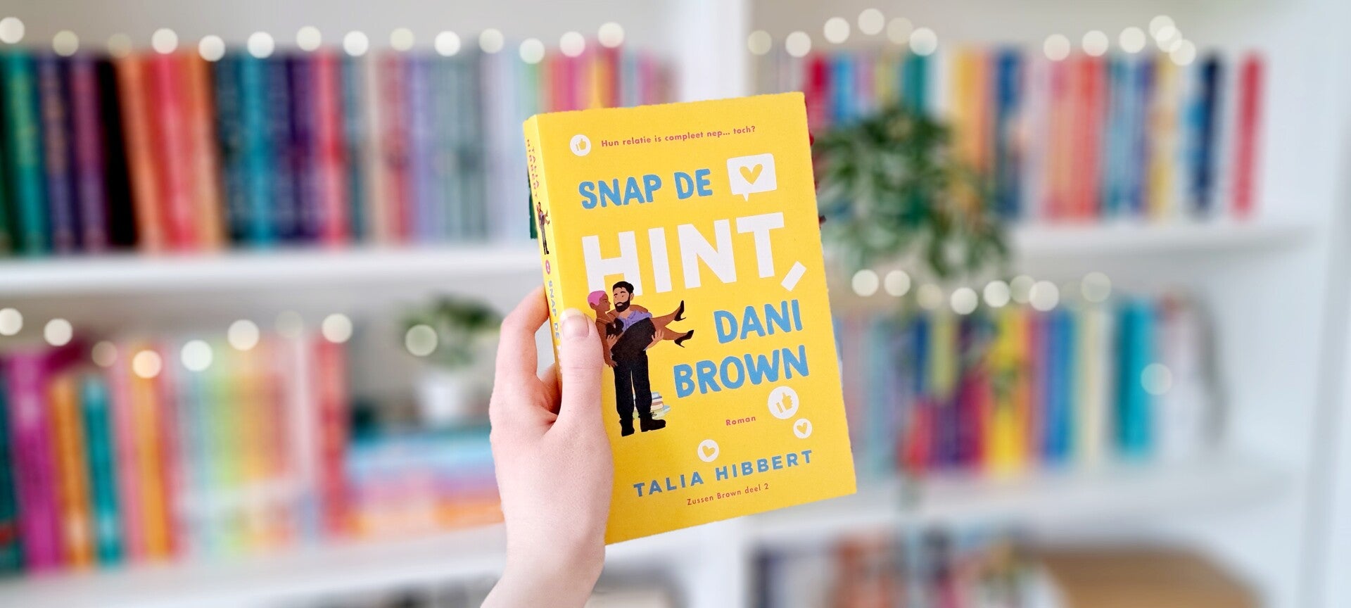 Een witte hand houdt 'Snap de hint, Dani Brown' deel twee van de Brown Sisters trilogie van Talia Hibbert omhoog voor een kleurrijke wazige boekenkast met lichtjes. Foto bij recensie door turnthepages.nl Rolstoelgebruiker, disabled bookstagrammer en queer