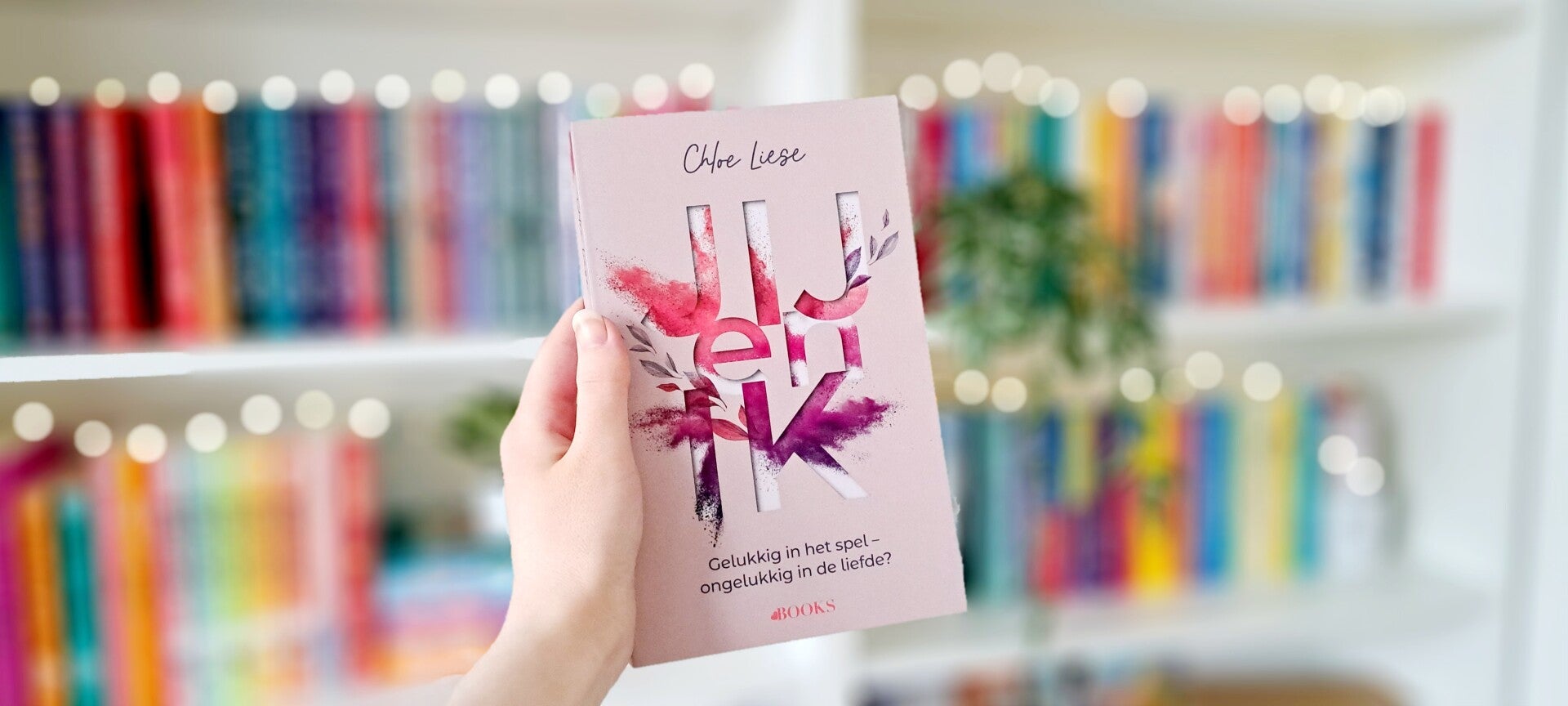 Een witte hand houdt 'Jij en ik' van Chloe Liese, deel 1 in de bergman brothers series/de bergman broers serie omhoog voor een kleurrijke wazige boekenkast met lichtjes. Foto bij recensie door turnthepages.nl Rolstoelgebruiker en disabled bookstagrammer e